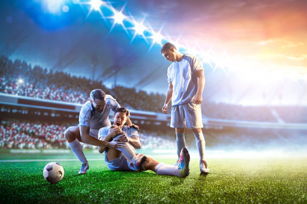 Football player helps injured player on a stadium field. Another player looks on. Sunset in background.