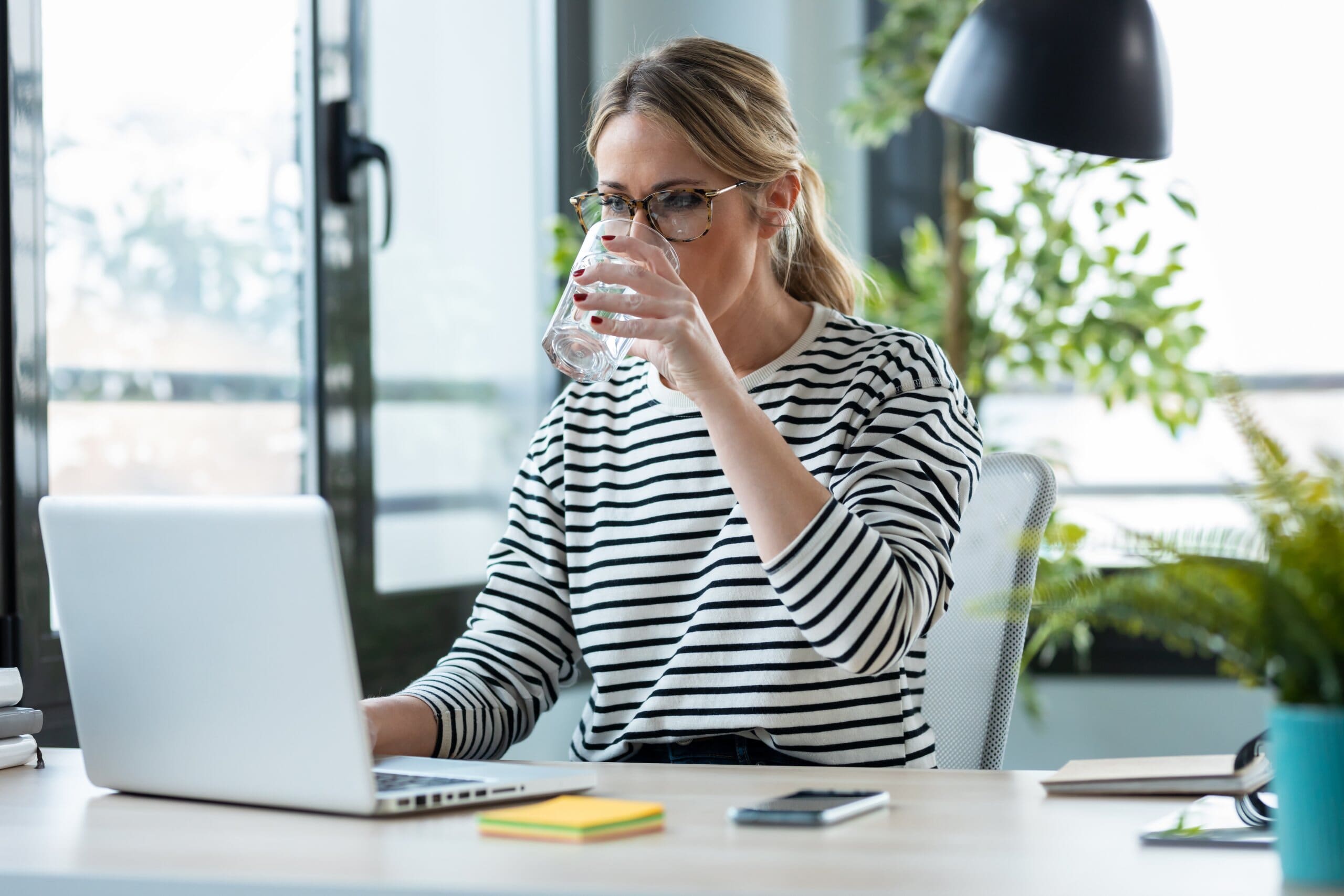 Woman sat at desk on laptop, drinking a glass of water.