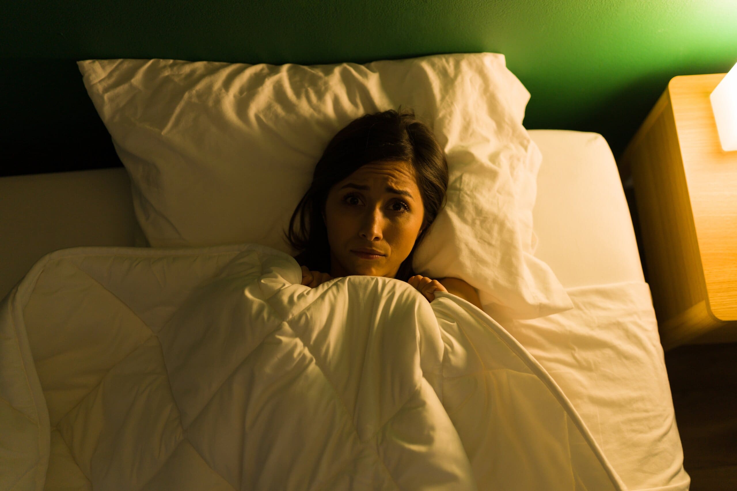 Woman laying in bed with duvet up to her chin, looking frightened.