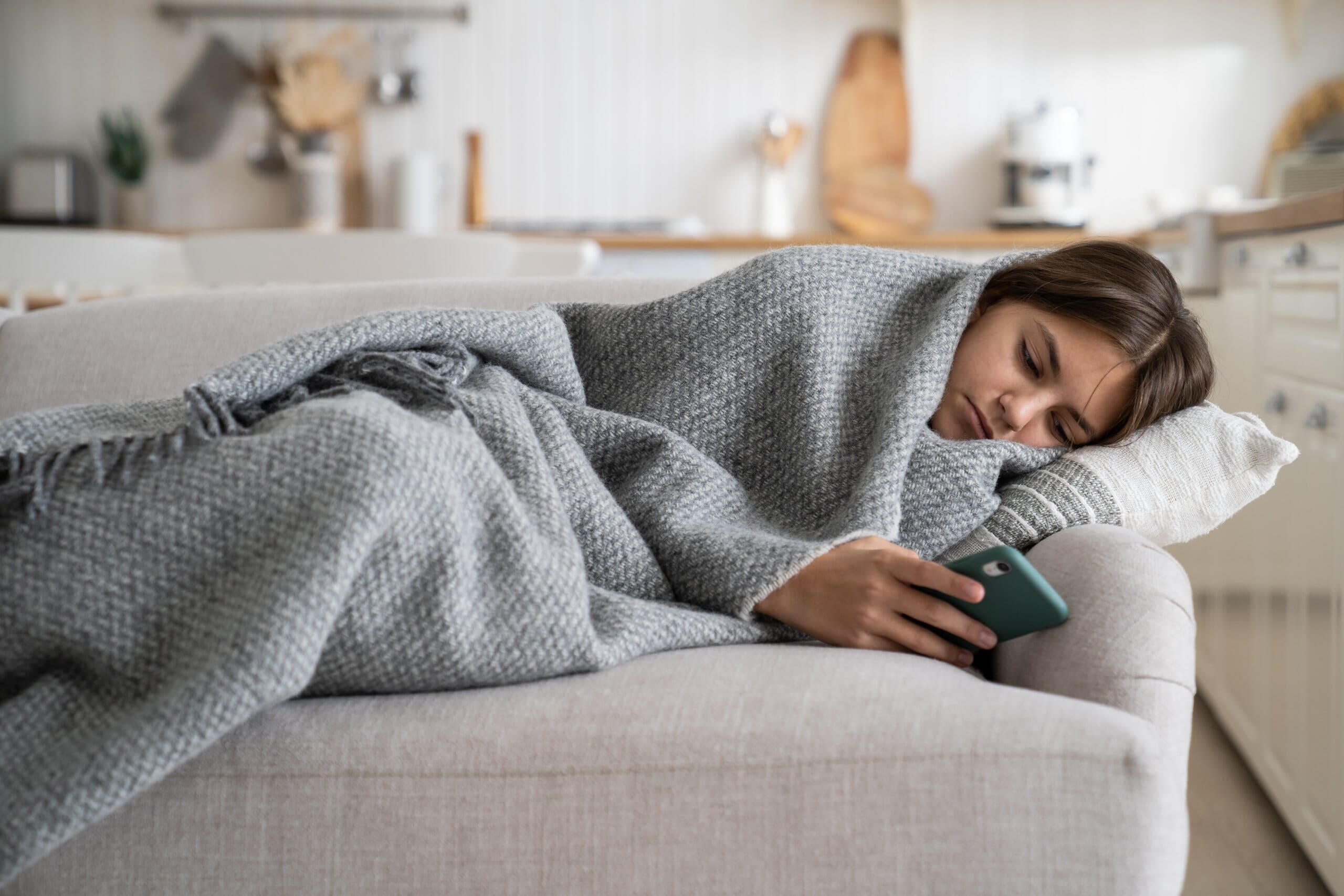 Girl looking depressed wrapped in a blanket on her phone on the sofa.