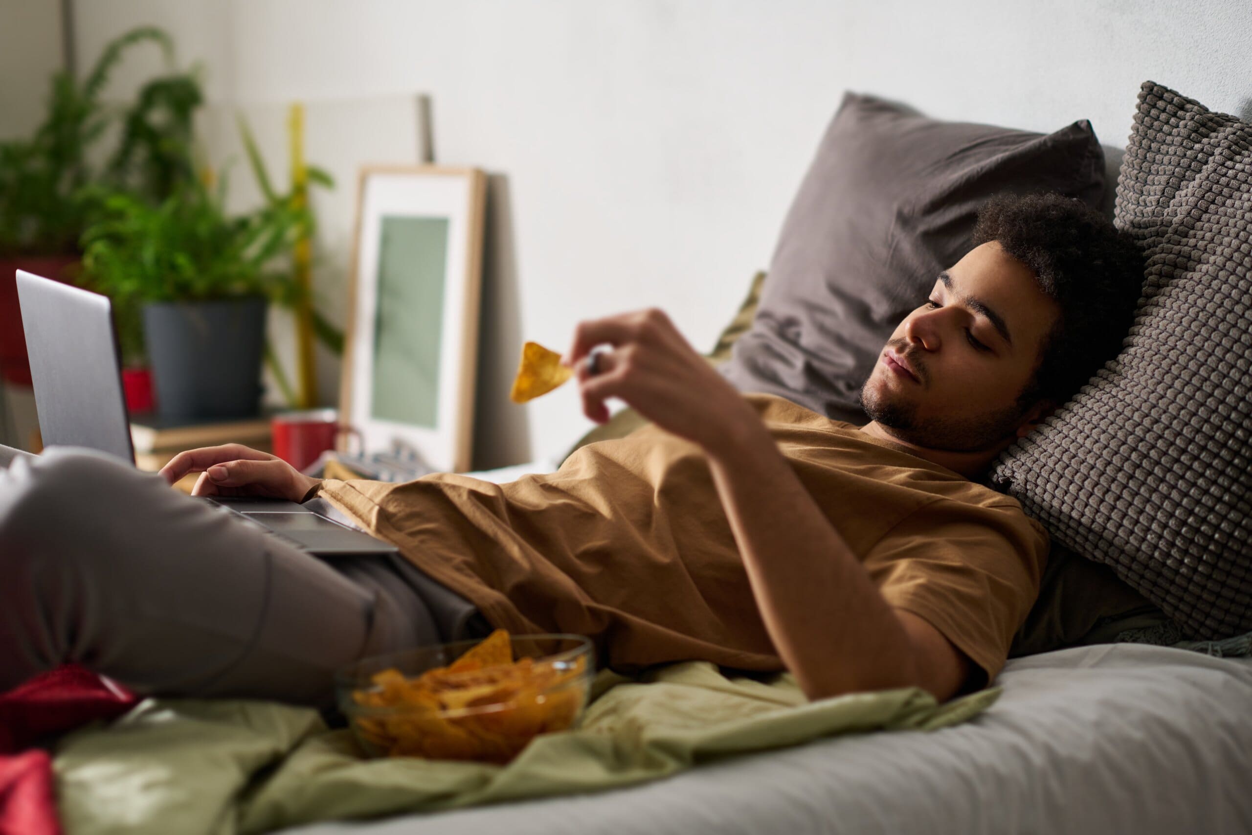 Lazy man laying in bed eating crisps on his laptop.