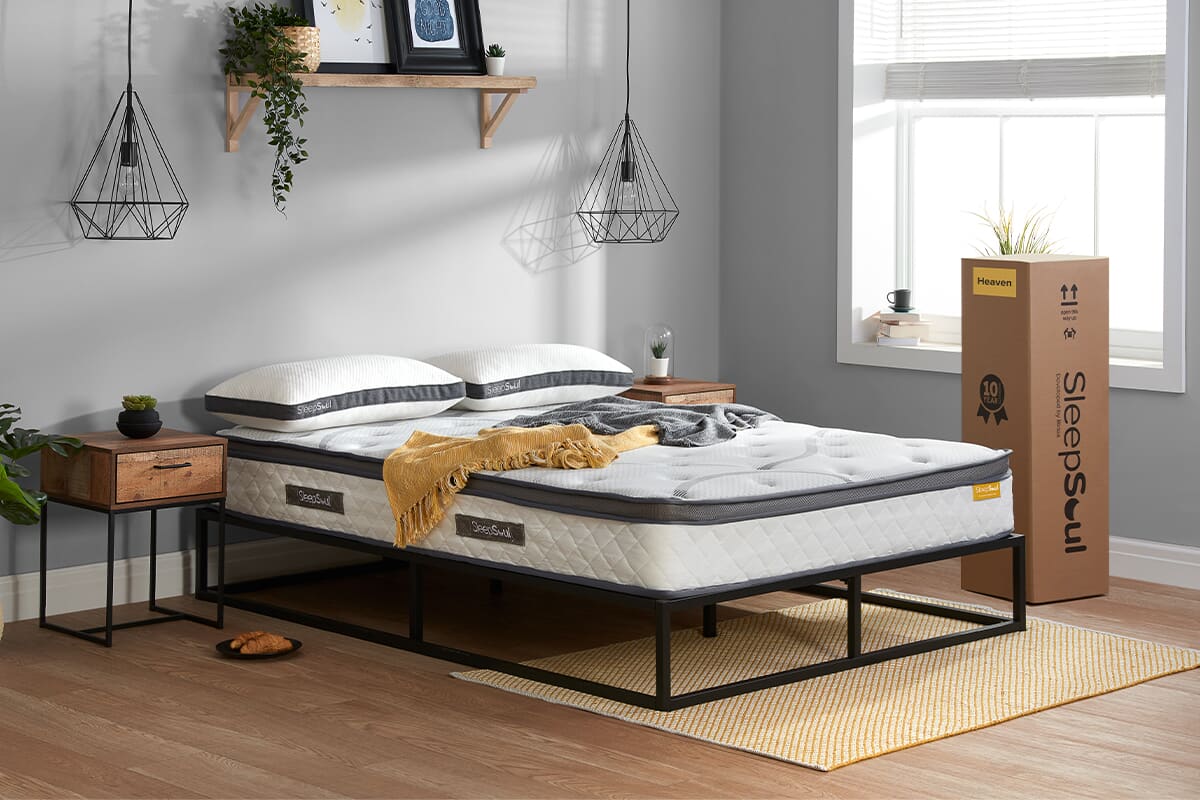 Lifestyle image of the SleepSoul heaven mattress on a metal bed frame in a modern bedroom. The mattress box is upright next to the bed.