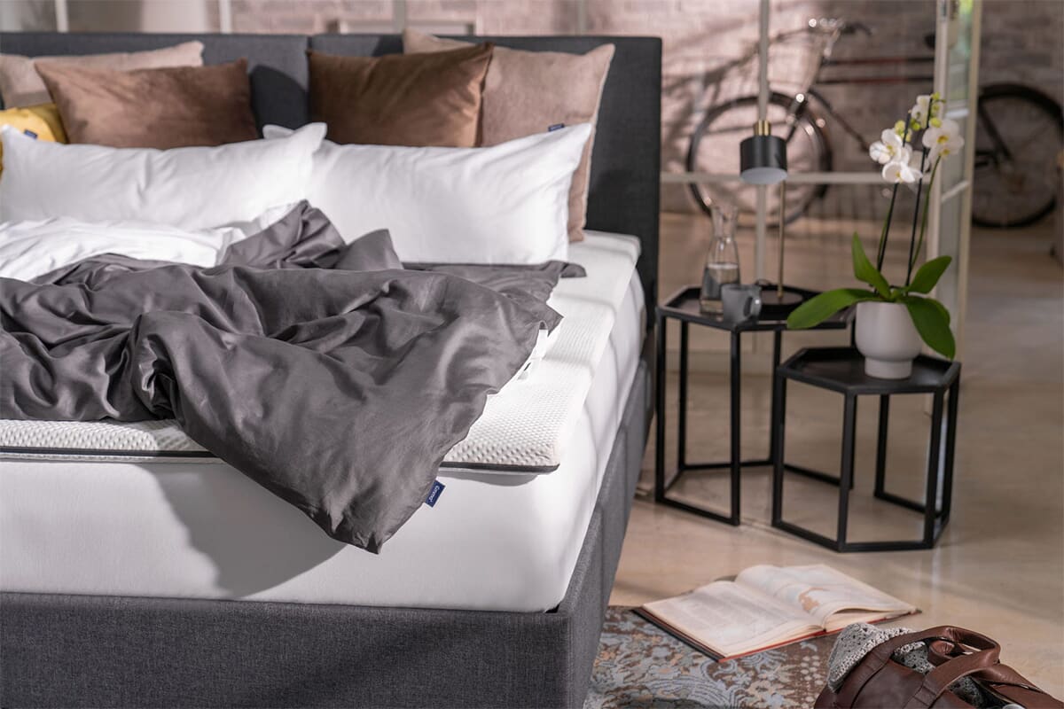 Image of a bed with the Emma Flip Topper on it, with messy cool bedding and bedside table.