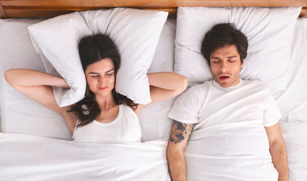 Top view image of couple in bed, man with mouth open and woman holding pillow to her ears.