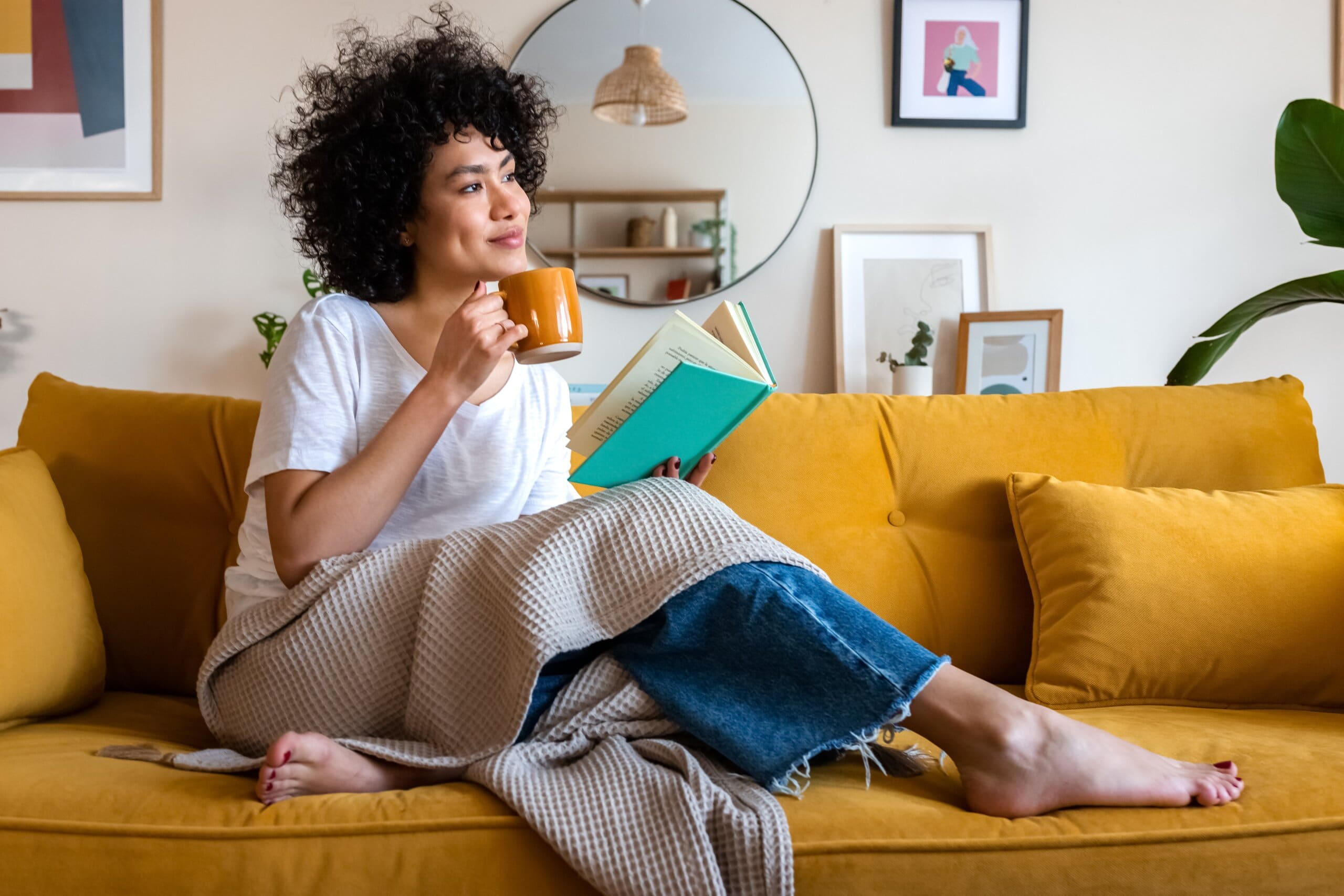 Relaxed woman enjoying a hot drink on a yellow sofa with a blanket.