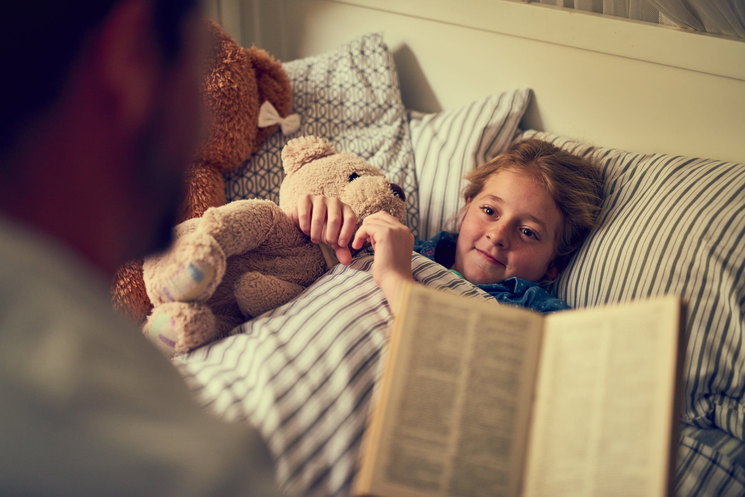 Father reading to a young girl snuggled up in bed with teddies.
