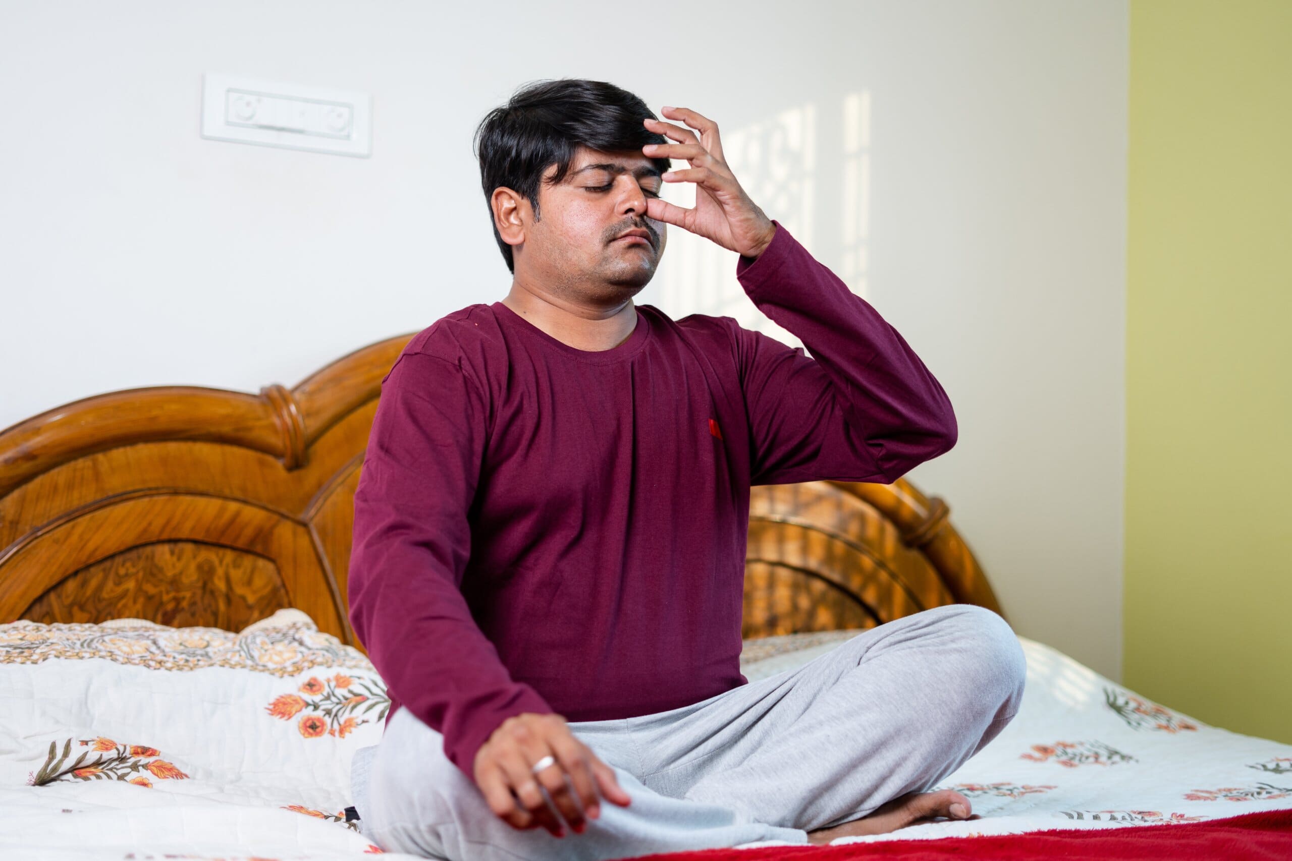 Man sitting cross legged on his bed, with one thumb pressed against the side of his nose and eyes closed, practicing breathing techniques.