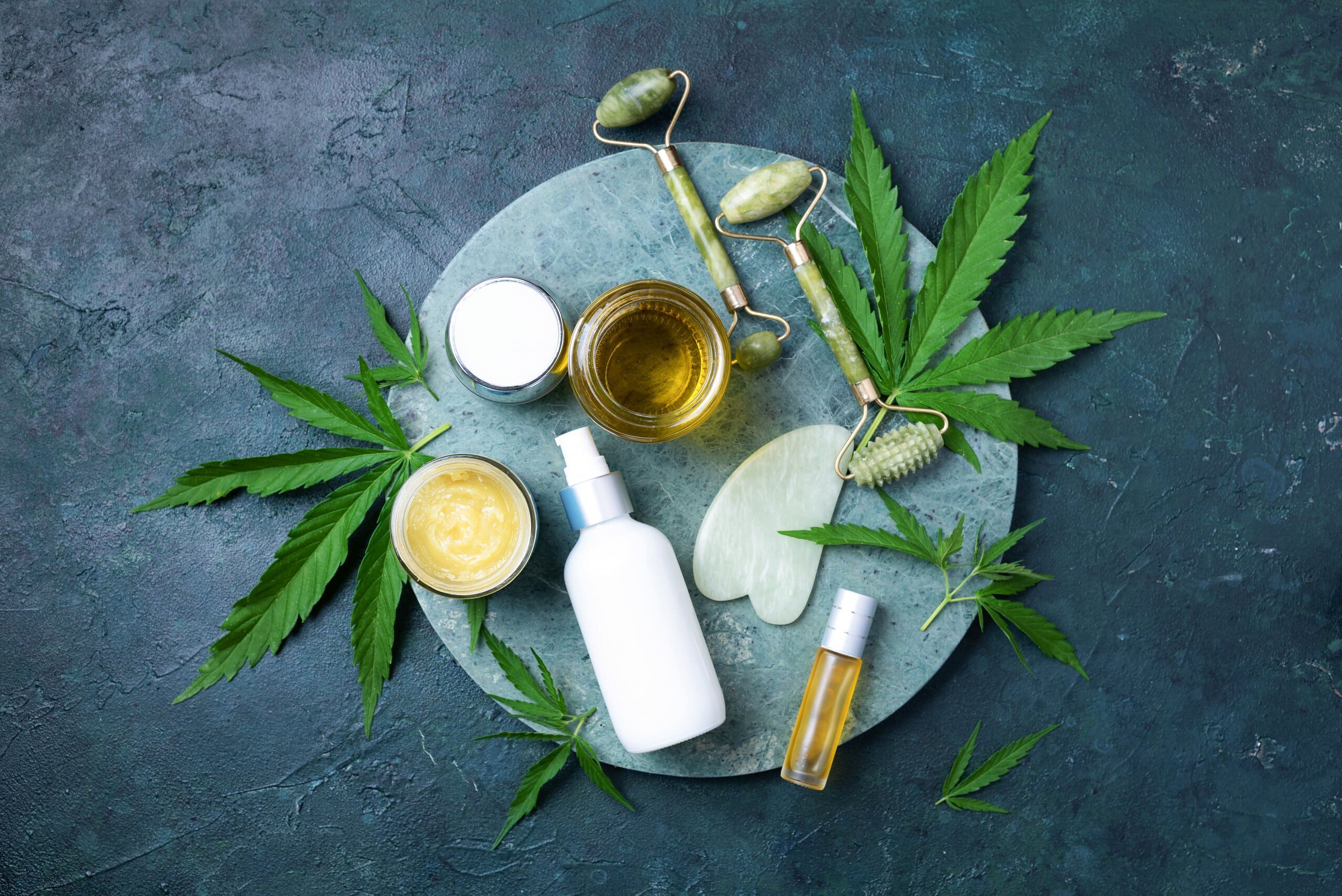 Hemp leaves and various creams, lotions and jade face roller on a circle tray.