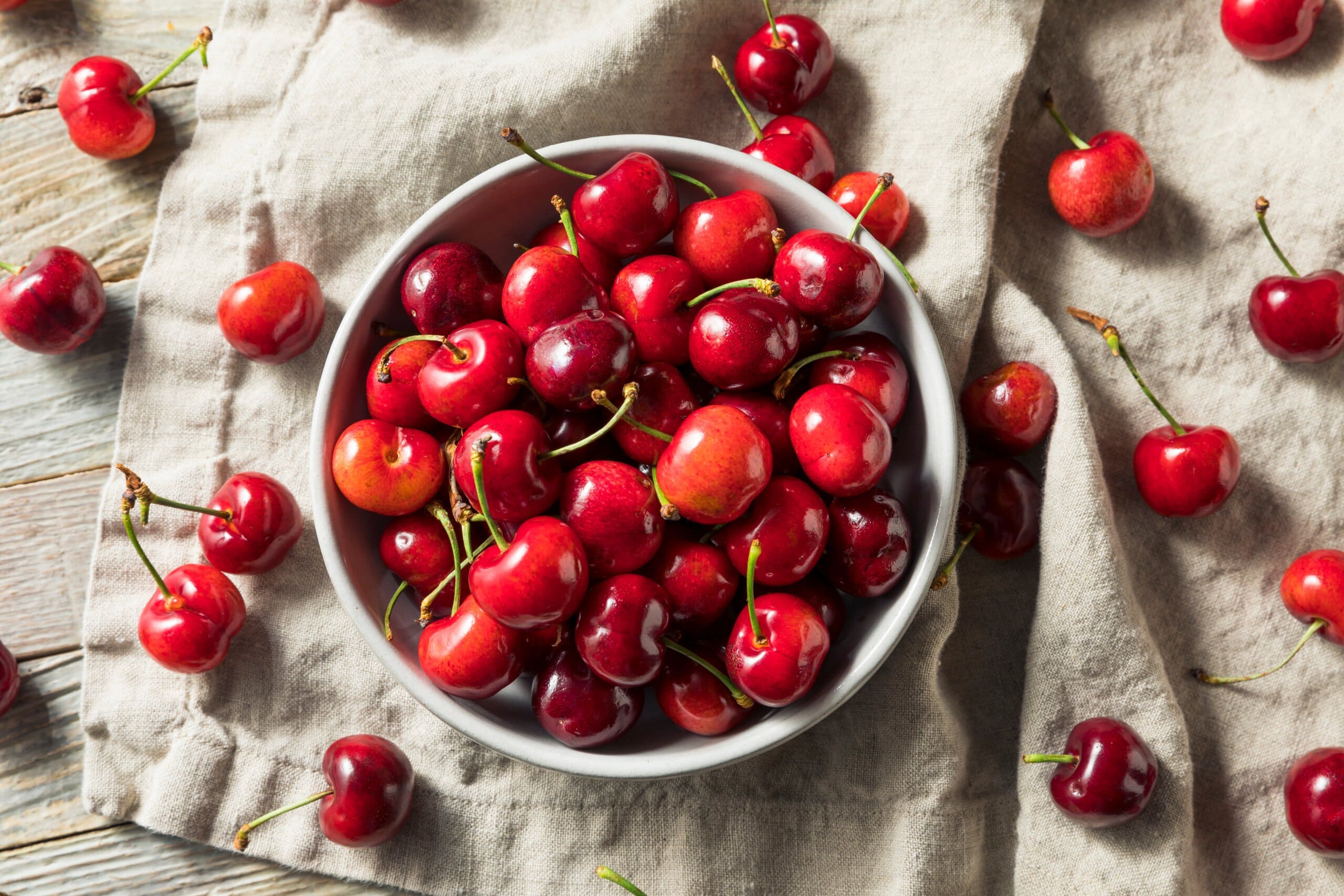 Cherries in a white bowl placed on a a table cloth with cherries around it.