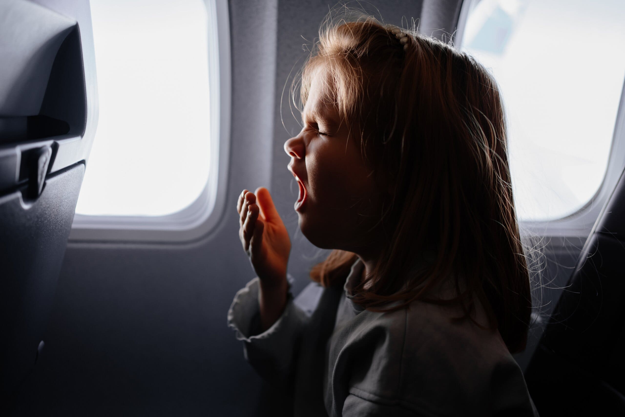 Young girl yawning into her hand in the window seat of a plane.