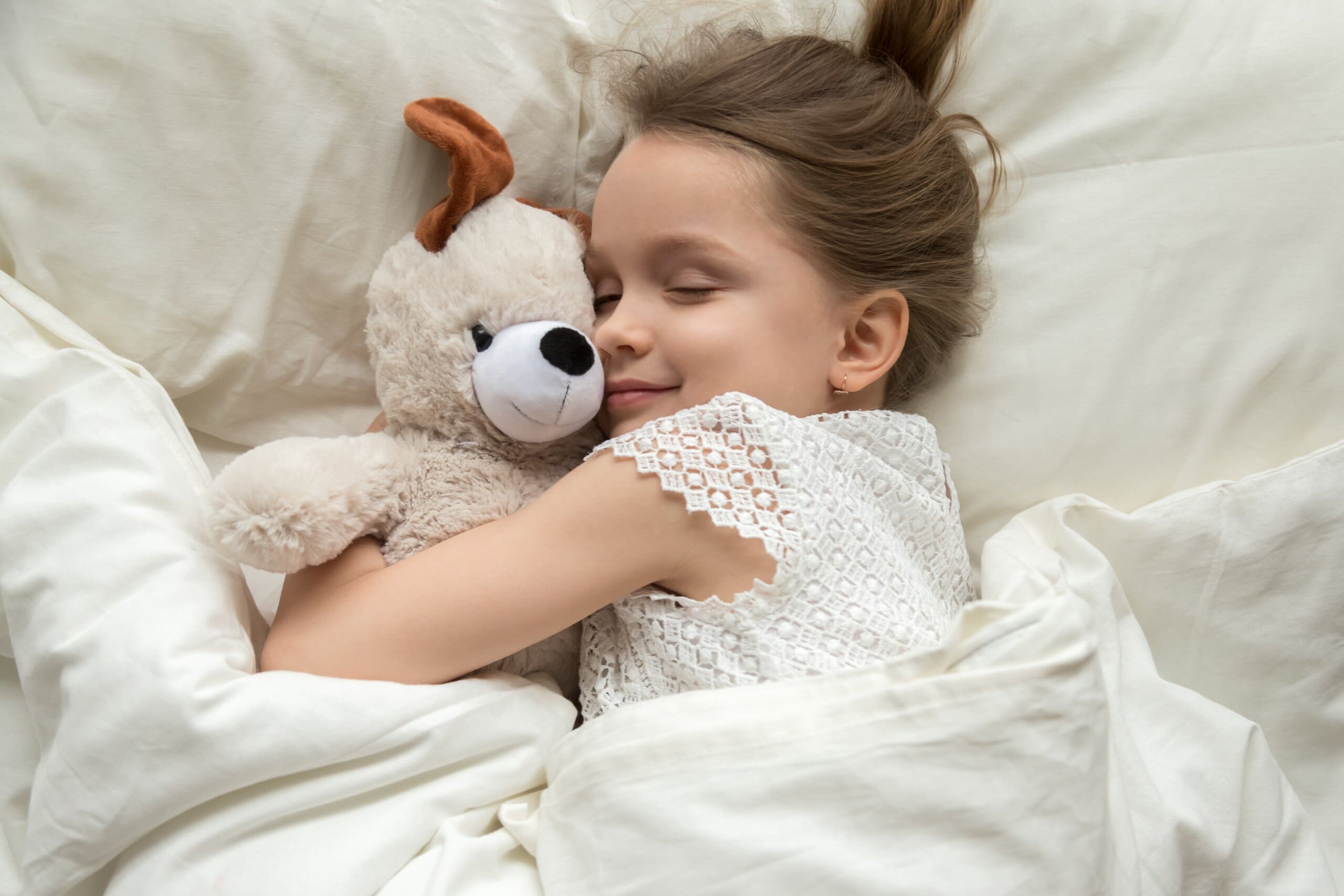 Little girl hugging her teddy bear smiling in bed with white covers over her.