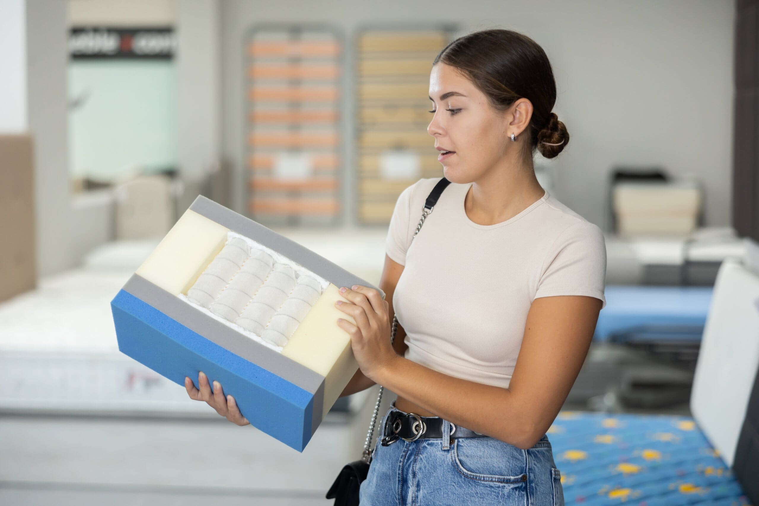 Young woman examines samples structure of spring mattress in furniture store. She studies different types of coils and their varying firmness, determined to find perfect fit.