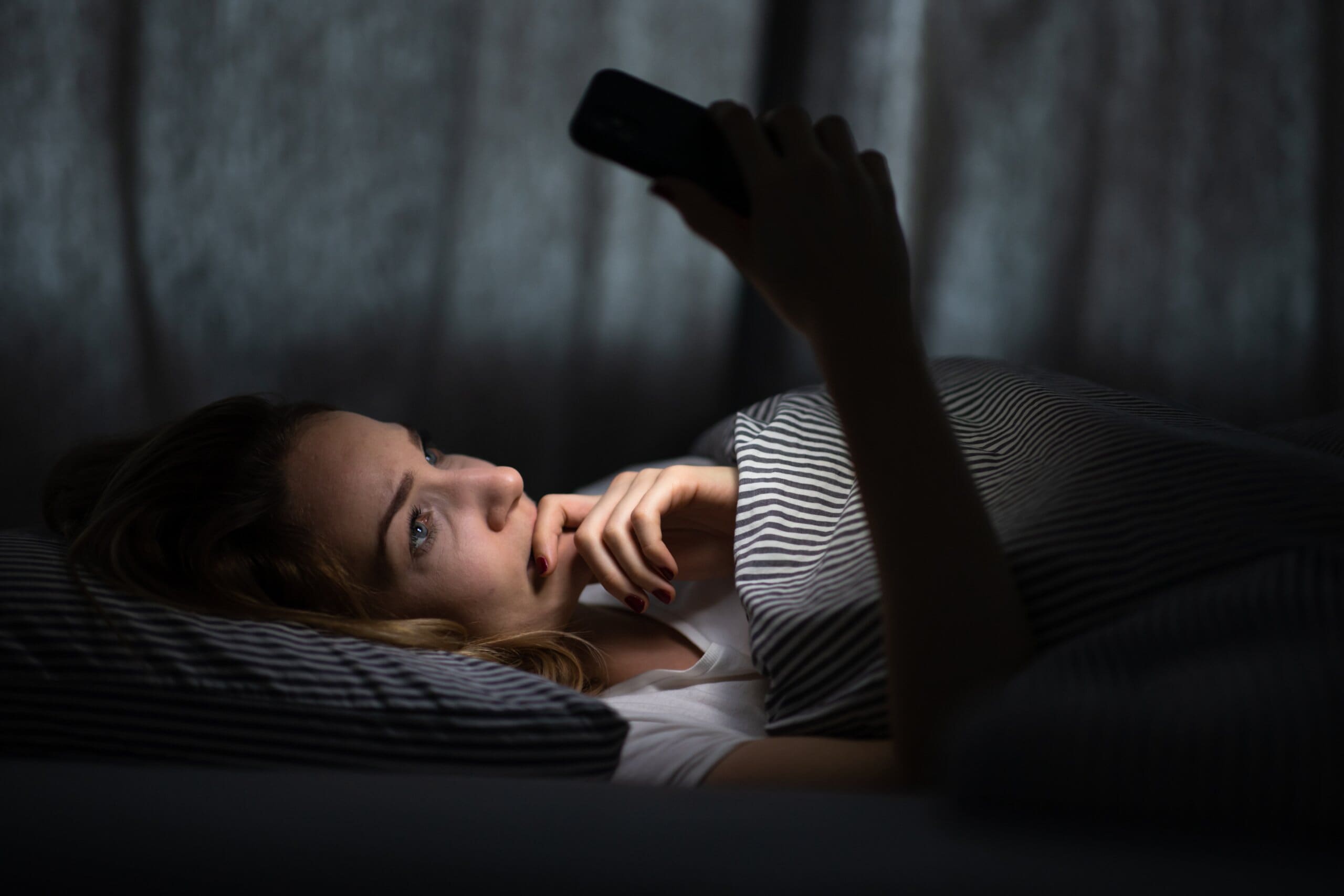 Young woman in bed holding a phone, tired and exhausted, blue light straining her eyes, messing up her circadian rhytm.