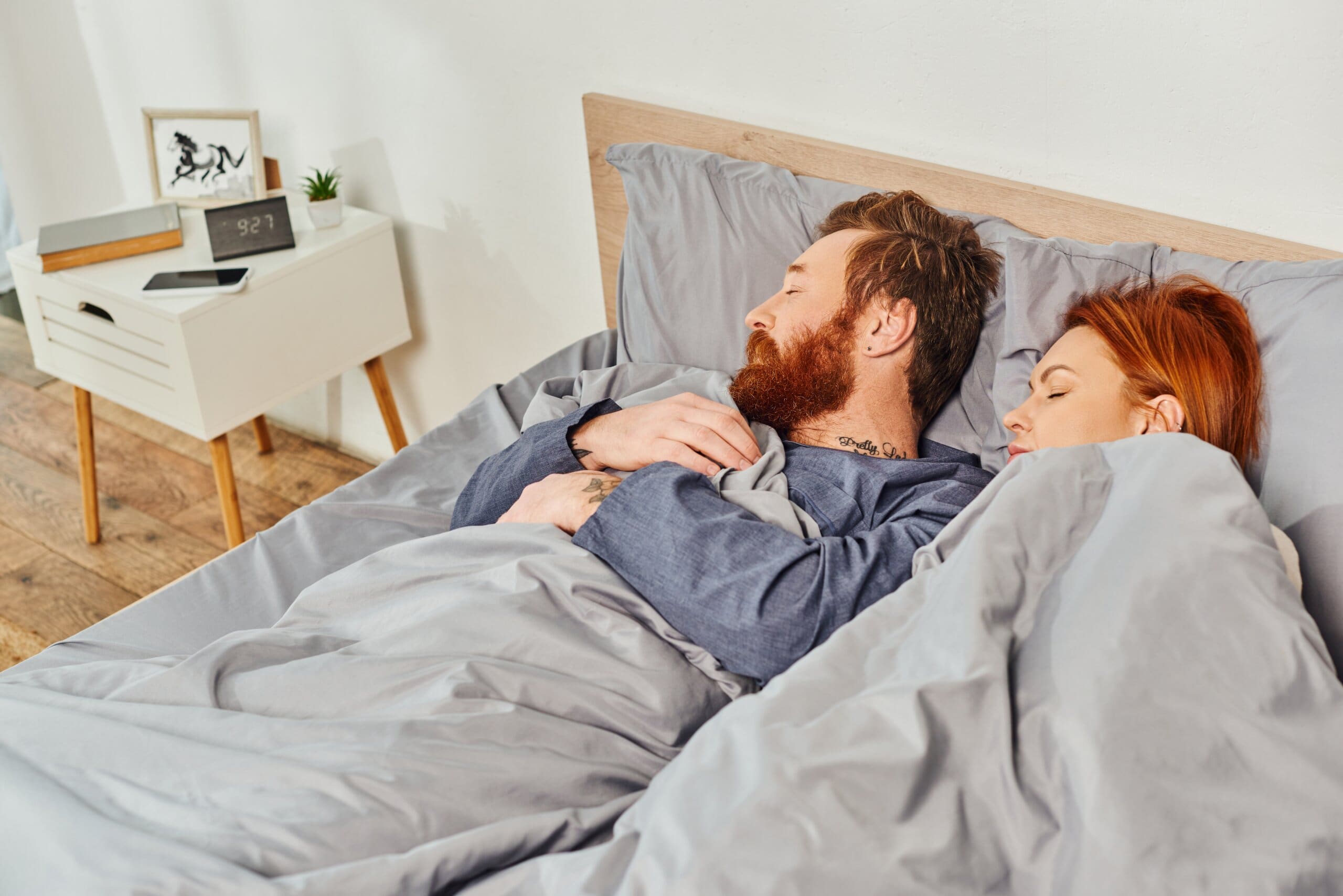 Couple asleep on their own sides of the bed next to the man's bedside table with his things on.