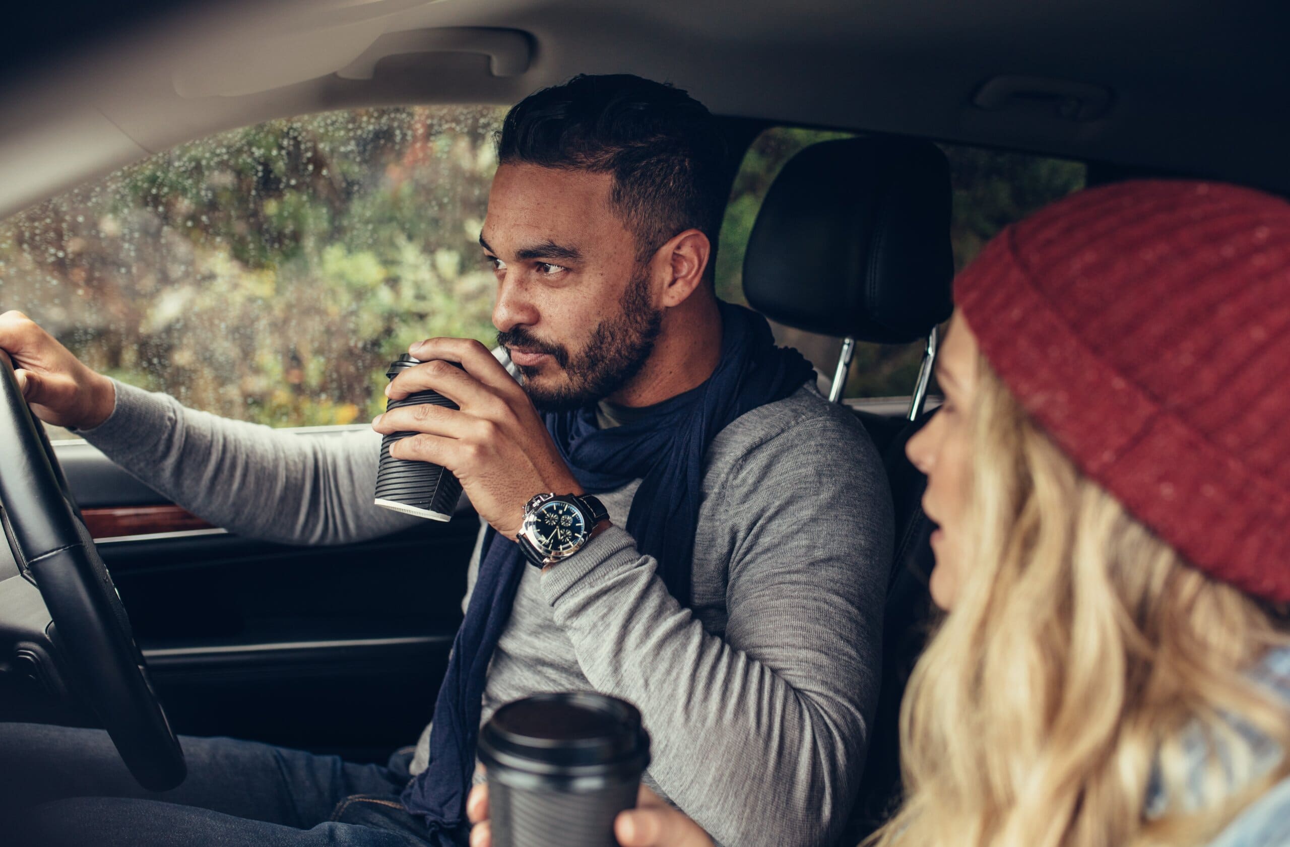 Couple drinking couple coffee in a car on a journey, wearing hats and scarves.