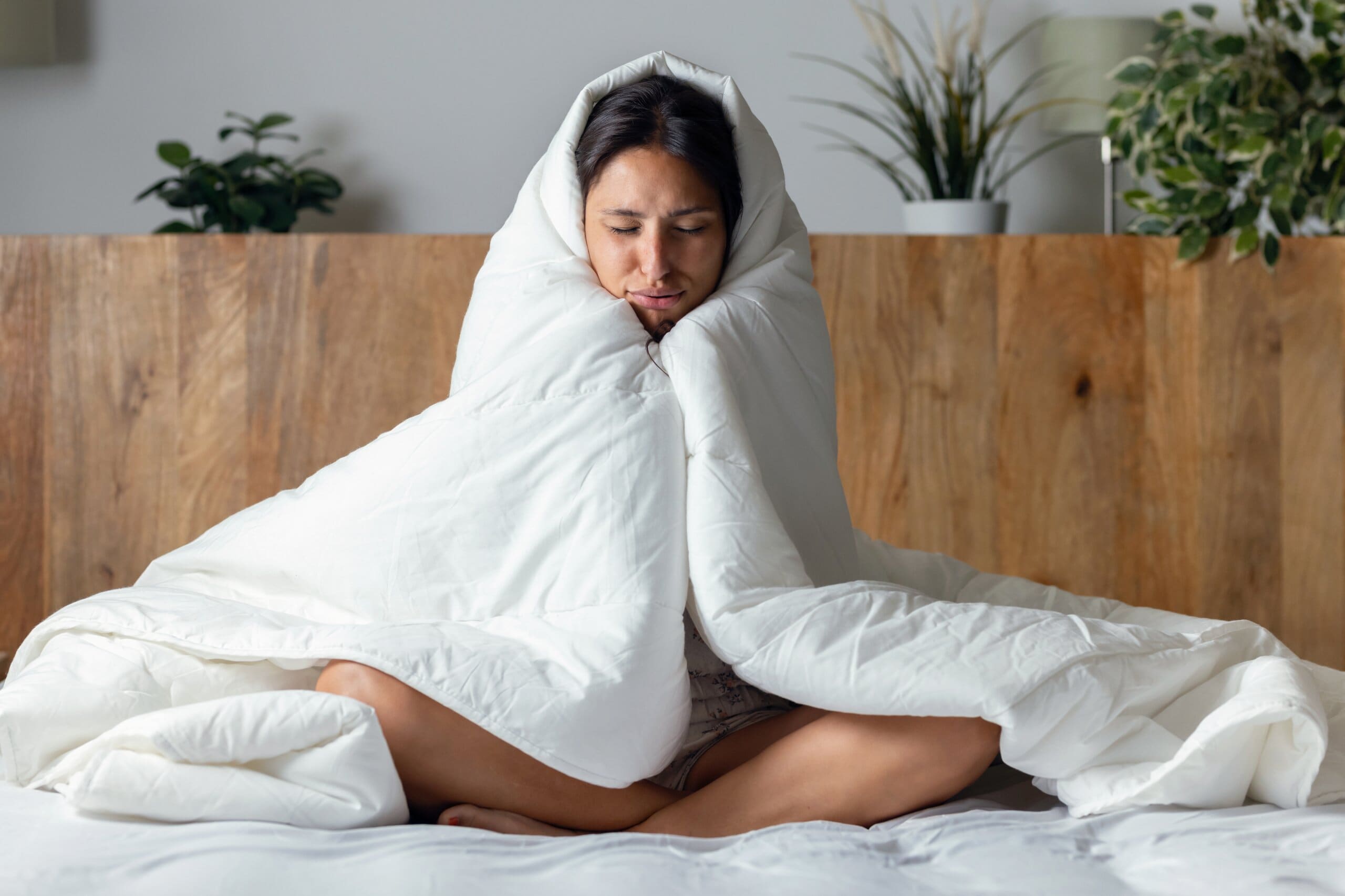 A woman sat cross-legged on the bed with her eyes closed, looking cold. She has a duvet wrapped tightly around her shoulders.