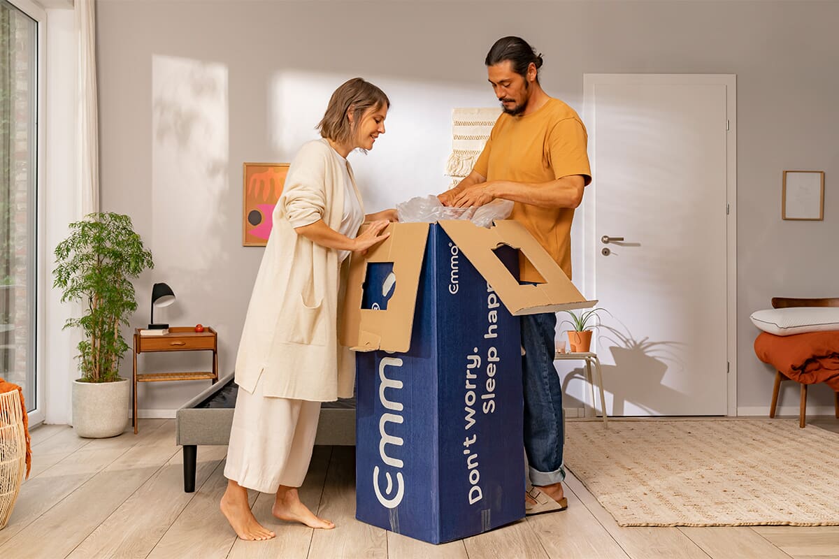 A man and woman unboxing an Emma mattress in a modern bedroom. They are peering into the upright box.