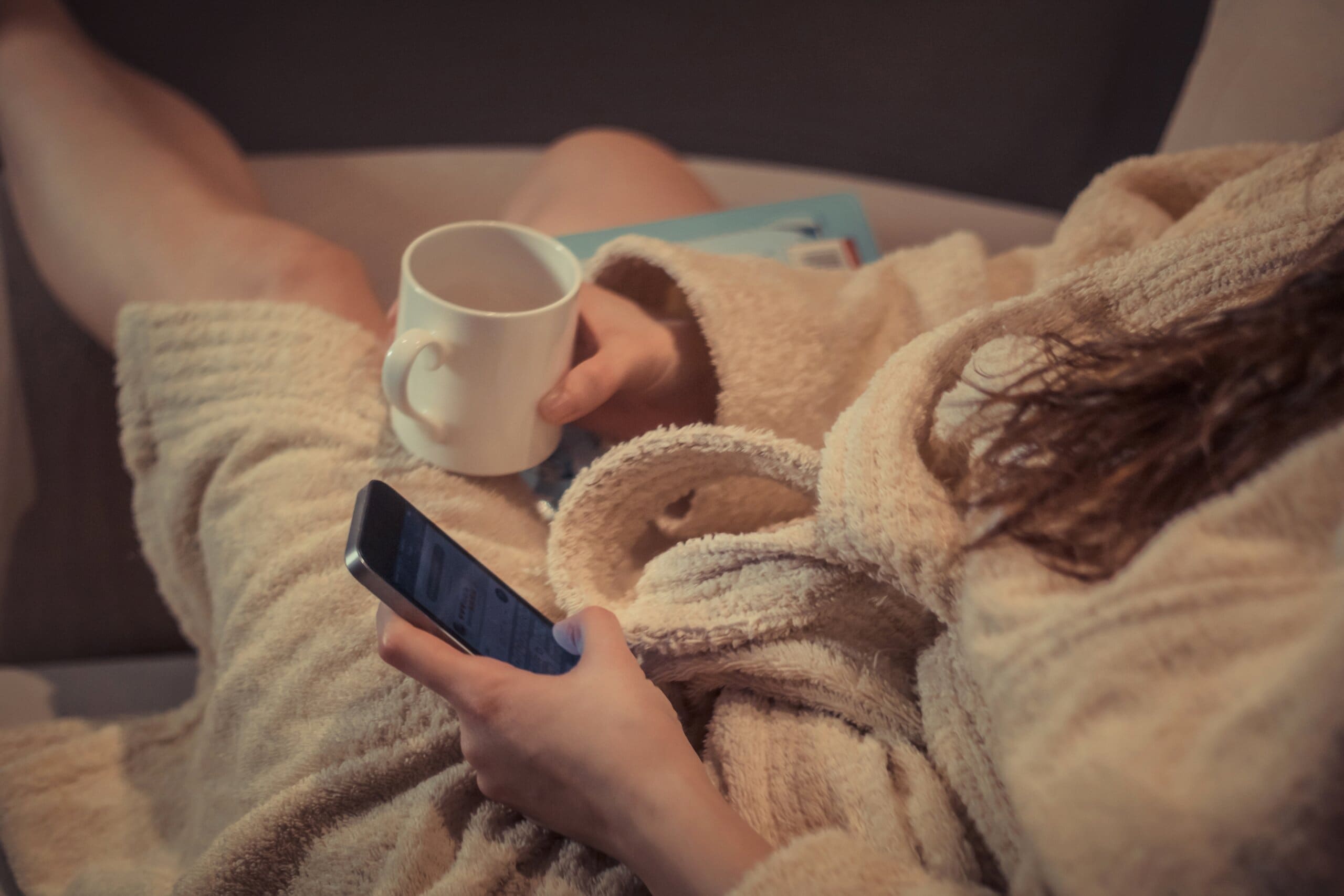 Close up of a woman in a bath robe after an evening shower, holding a mug and mobile phone, relaxing.