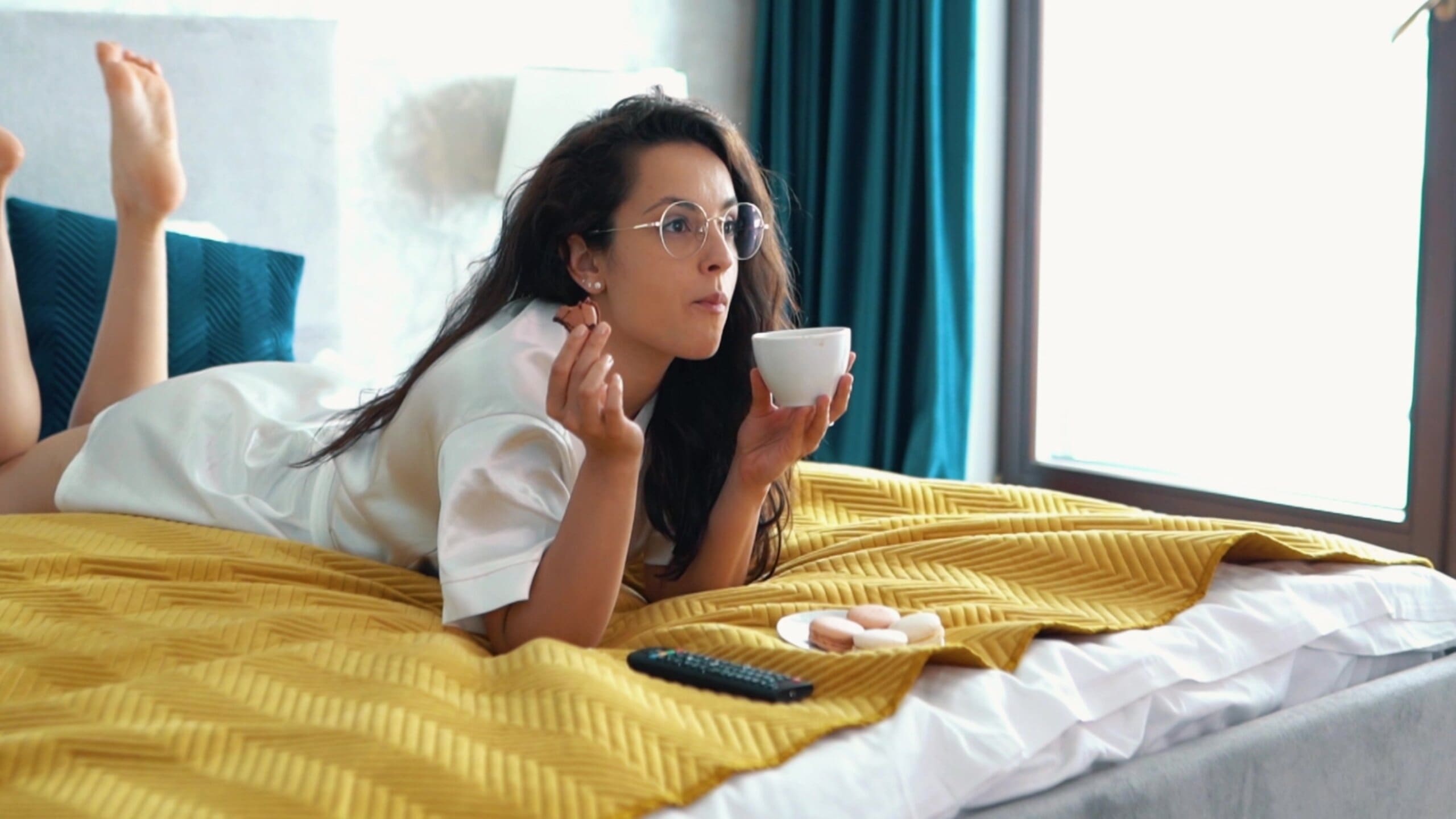 Woman laying on bed eating treats and drinking a hot drink.