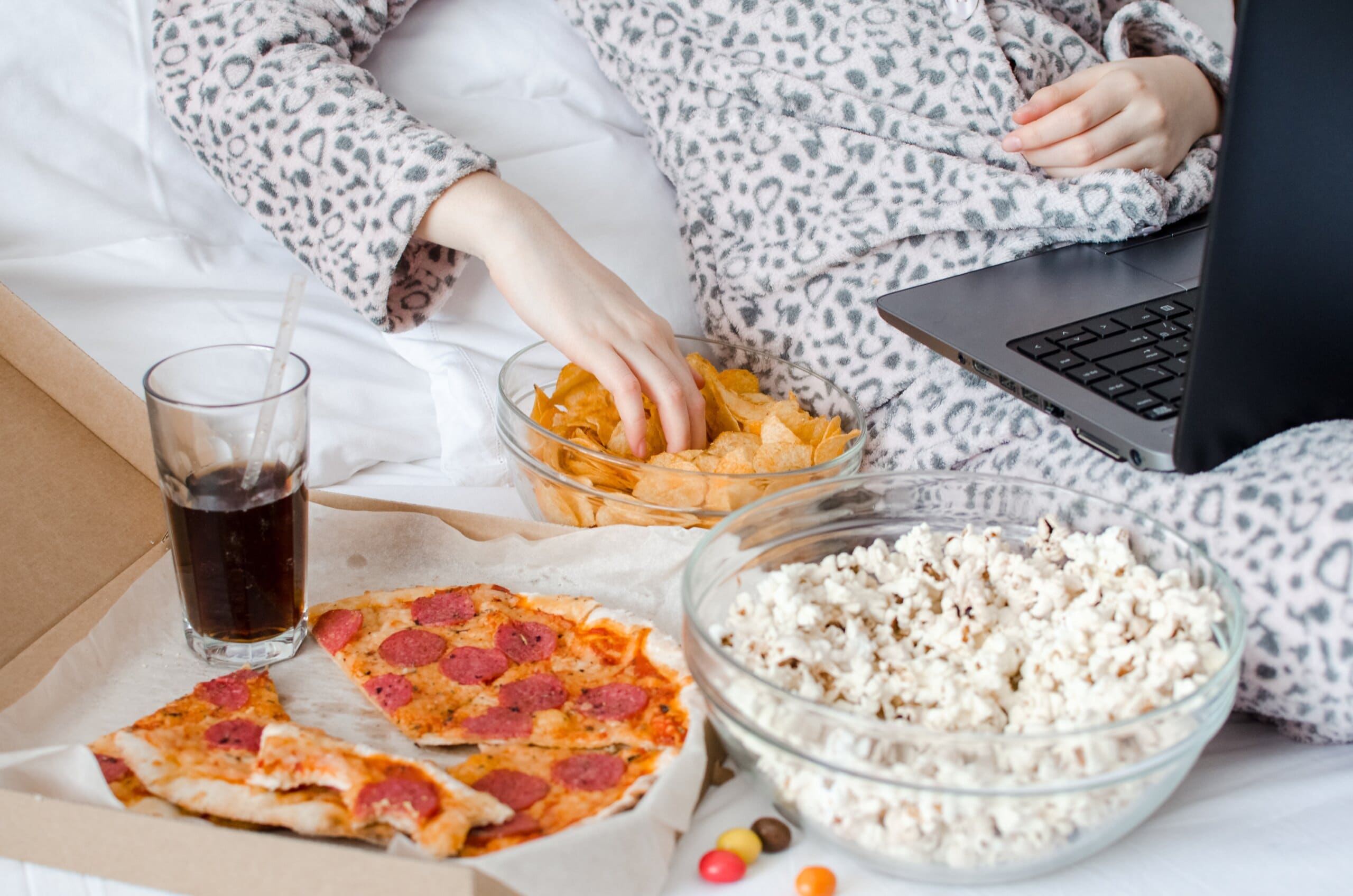Cropped image of woman laying in bed on laptop, with pizza, popcorn, drink, and crisps next to her.