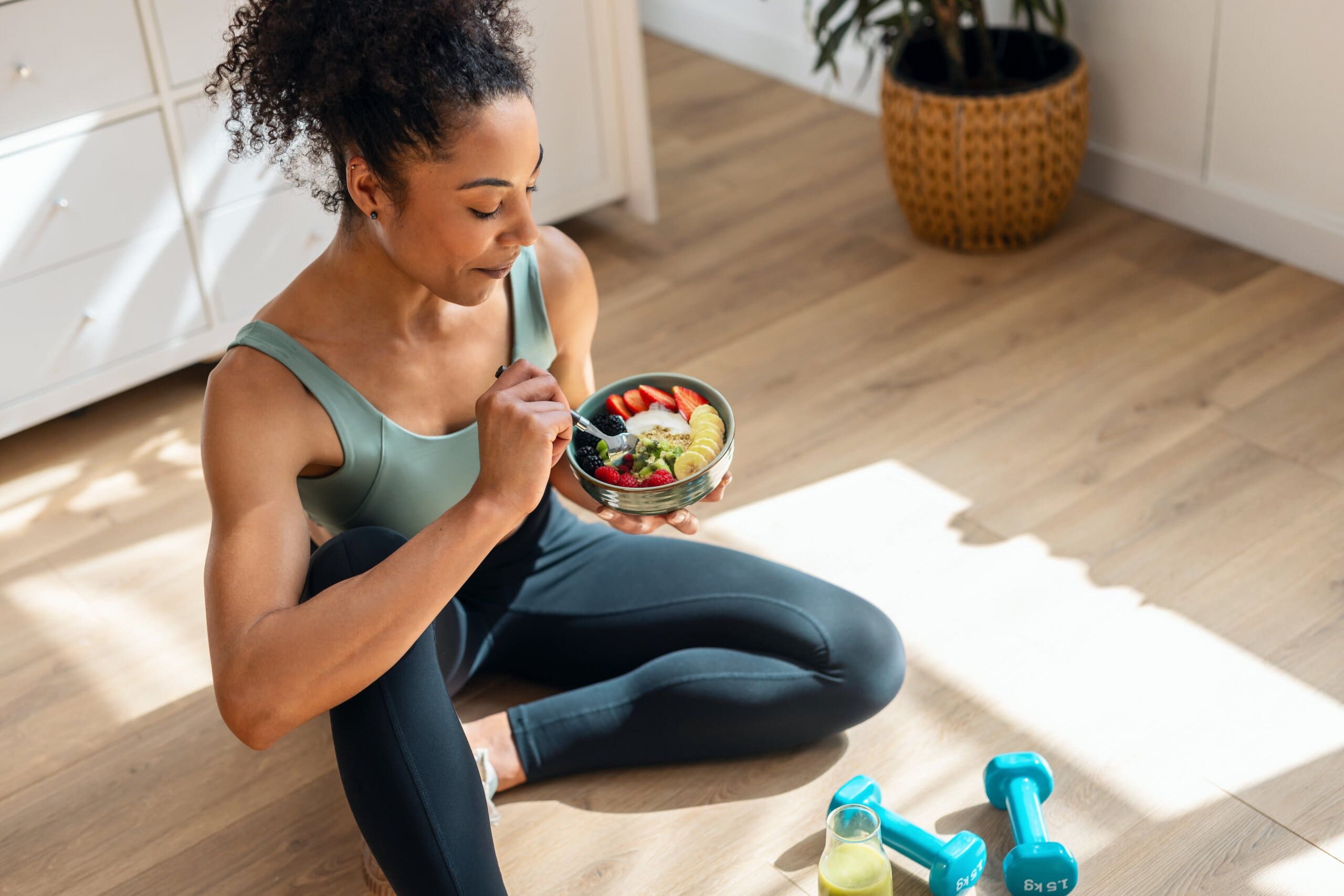 Woman in gym wear eating a bowl of fruit on the floor with weights next to her.