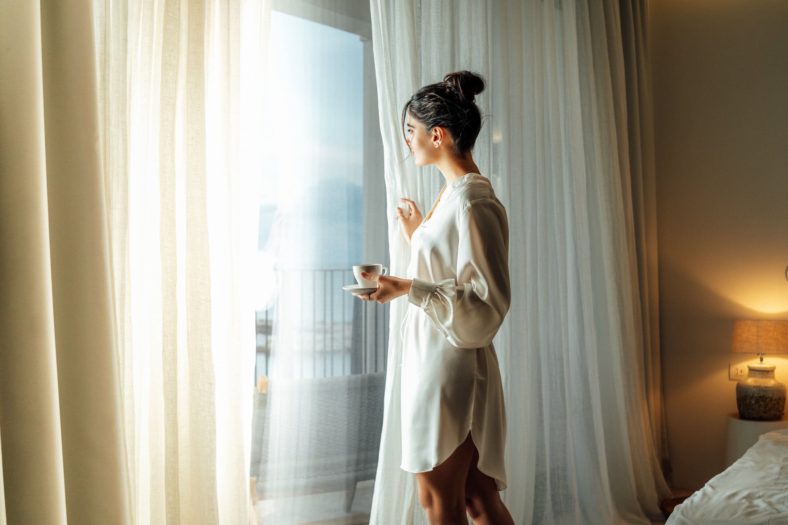 Young woman wearing white bathrobe opening curtains in luxury hotel room and enjoying sun in the morning while having her cup of coffee.