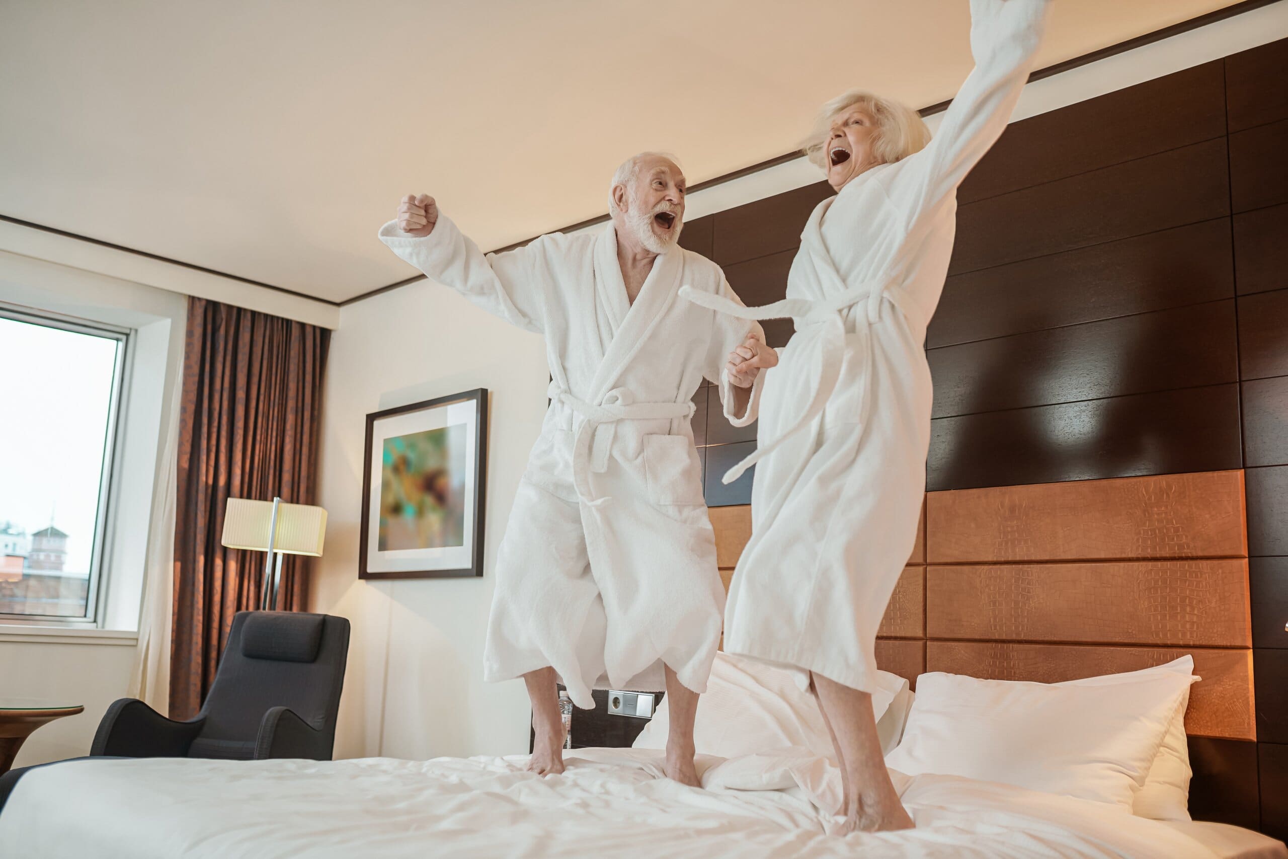 Senior couple in white dressing downs holding hands and jumping on a bed.
