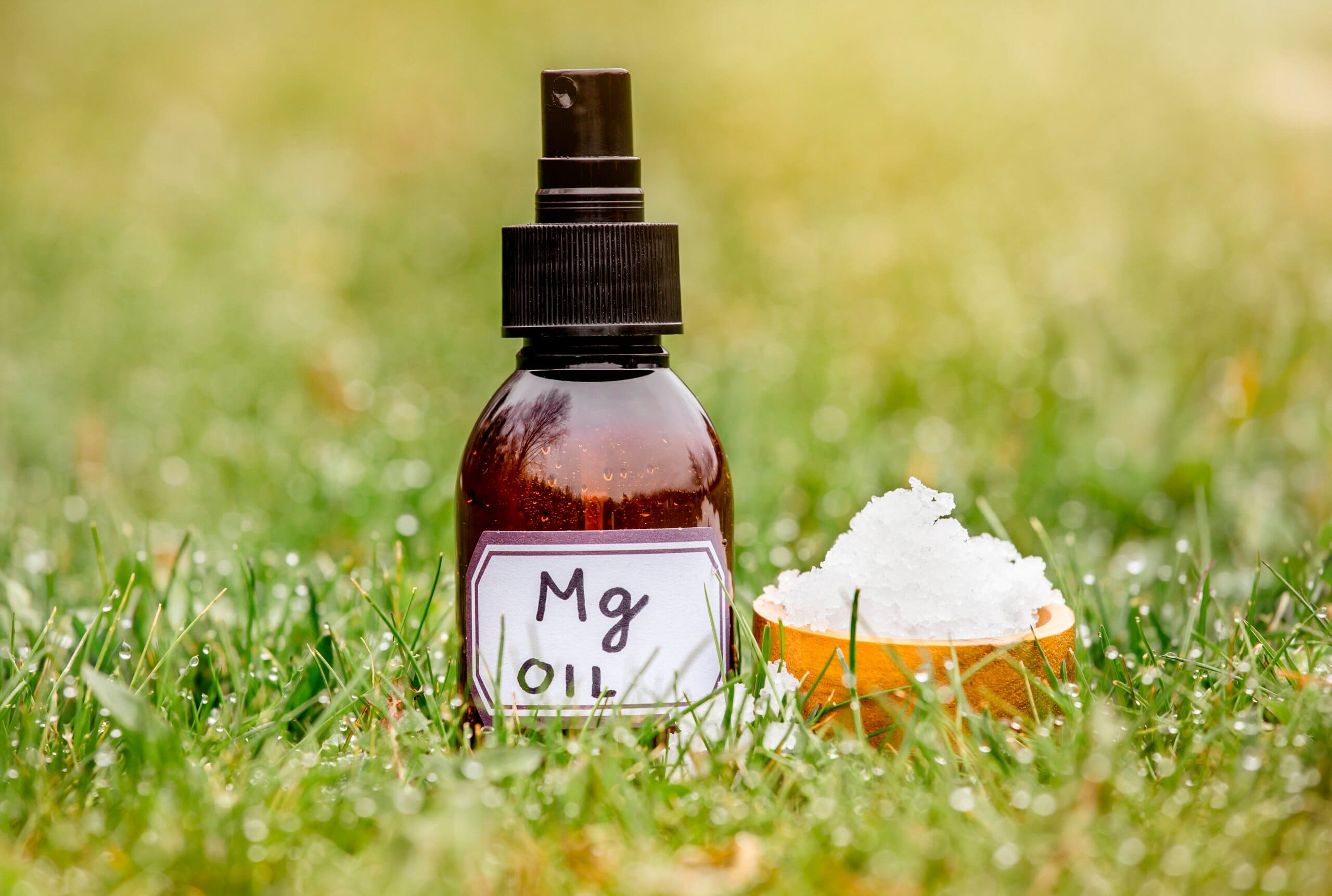 A spray bottle of magnesium oil next to a small wooden bowl of magnesium flakes on grass.
