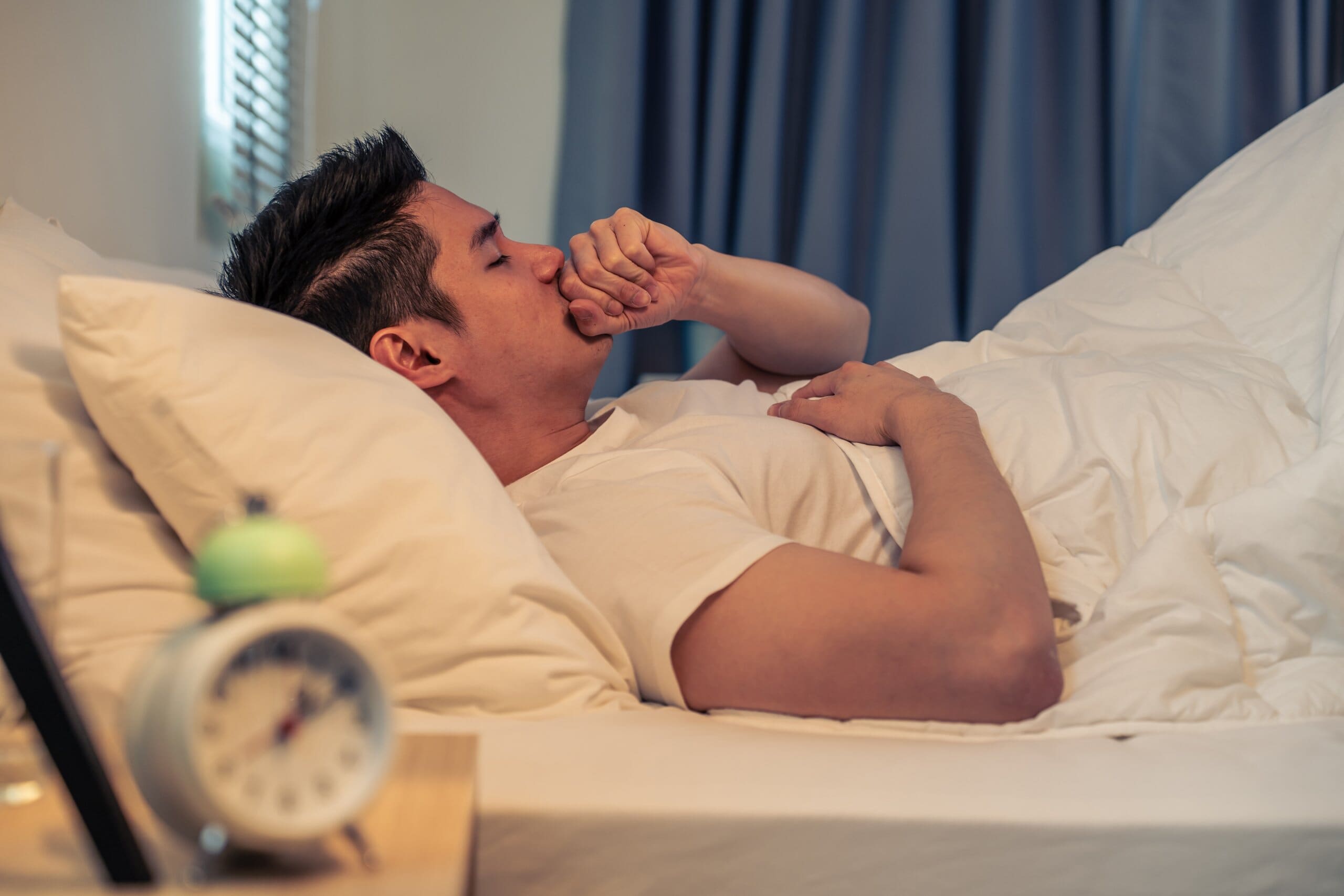 Man laying in bed, holding fist up to his mouth as he coughs.