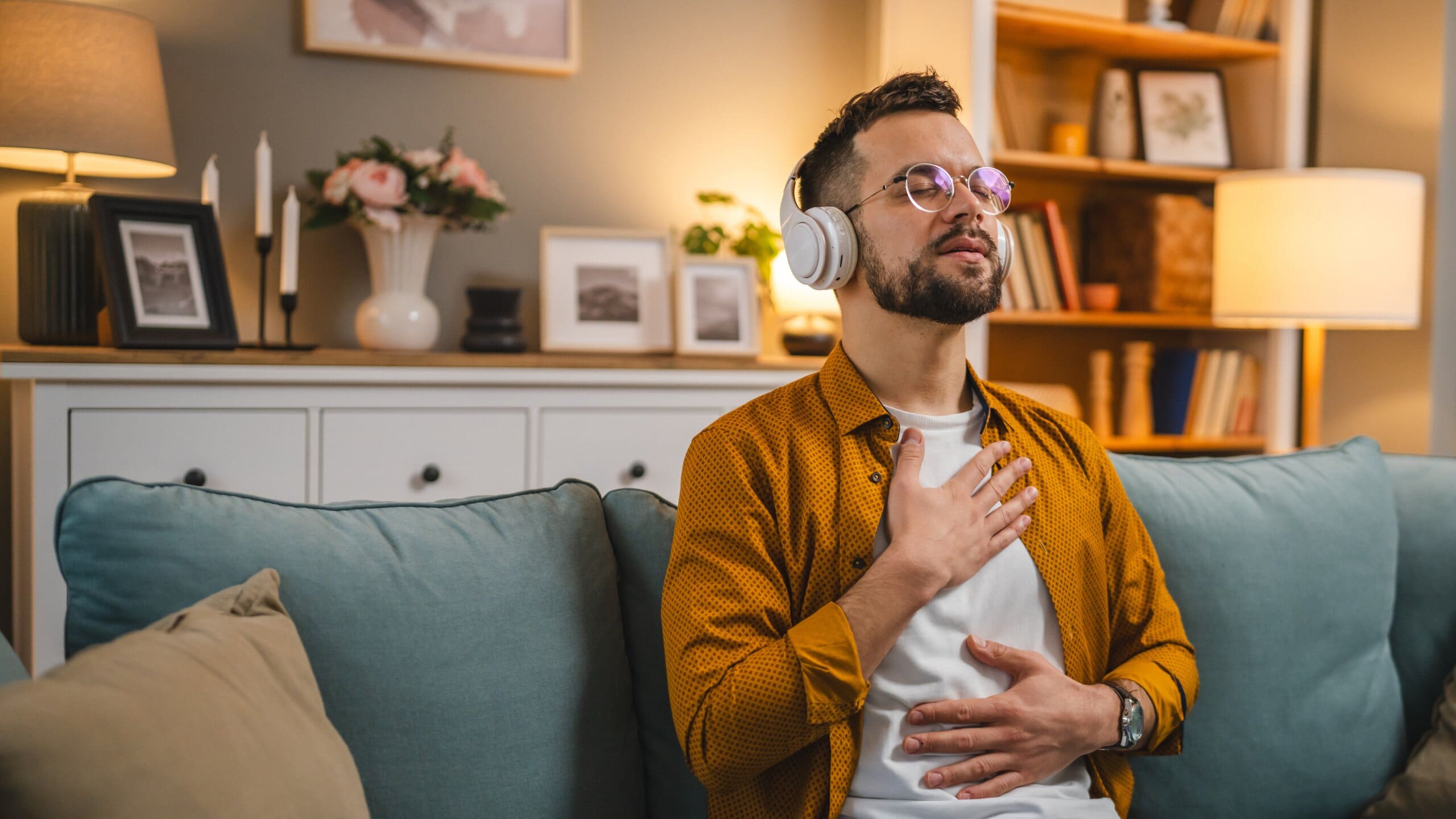 Man sitting on sofa with headphones on doing guided meditation, touching his chest.