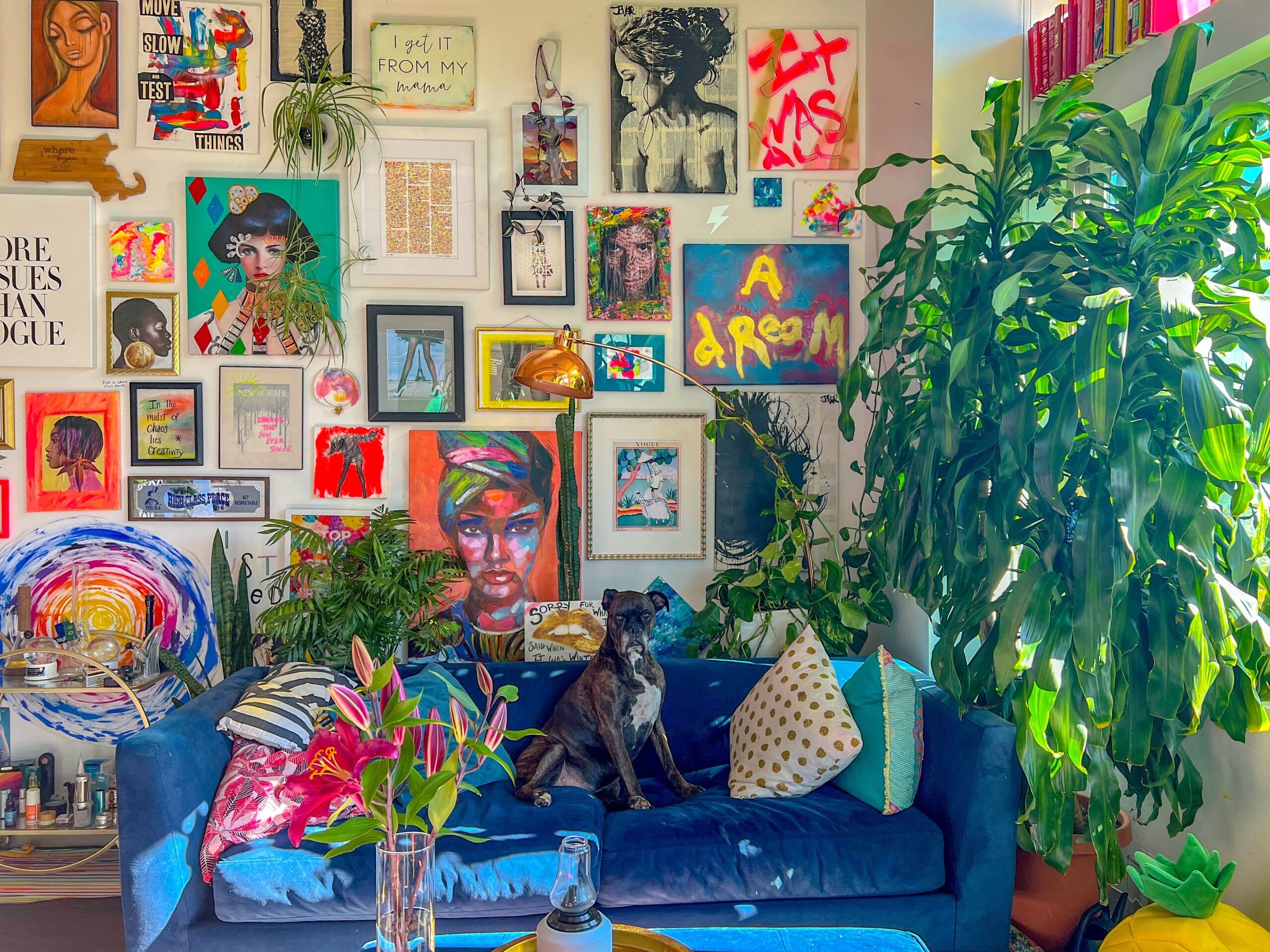 A maximalist bedroom wall covered in framed prints and artwork. A blue sofa with a dog sitting on it is in front of the wall.
