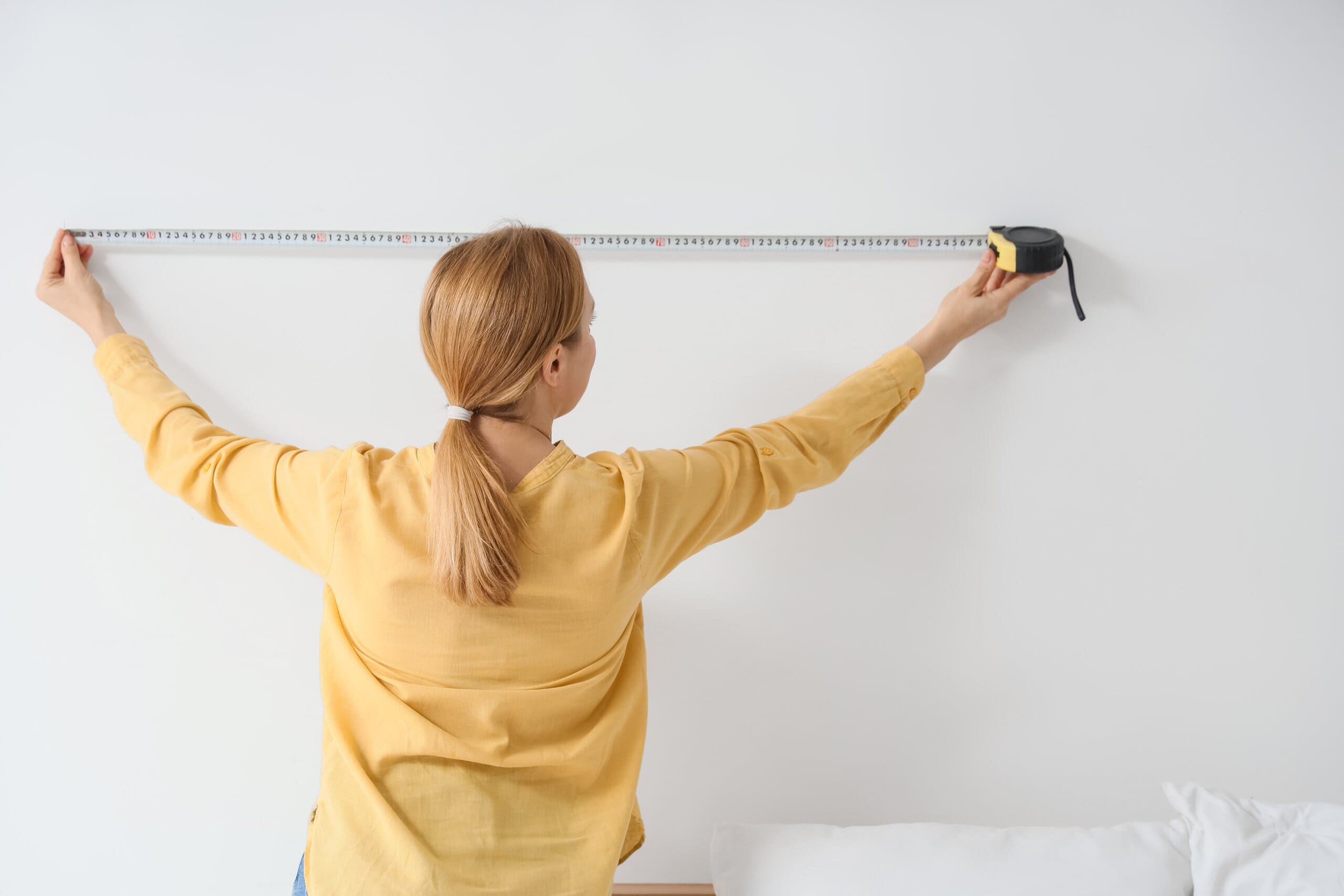 Woman in a yellow top with her back to the camera, holding up a tape measure to the wall.