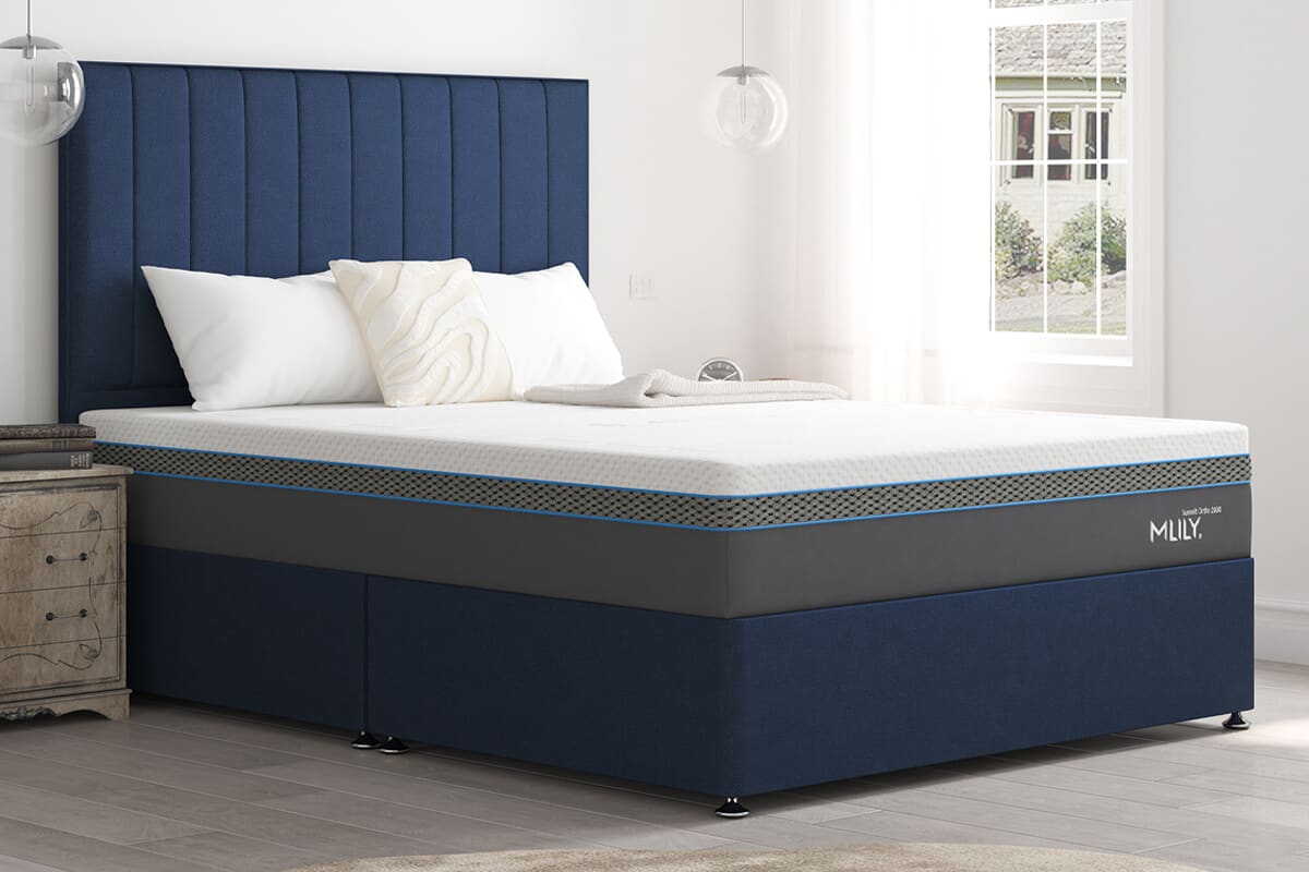 Side angled image of the MLILY Summit mattress on a dark blue divan bed in a brightly lit bedroom.