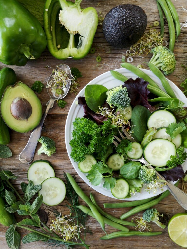 A green salad in a bowl surrounded by various other green vegetables on a wooden worktop.
