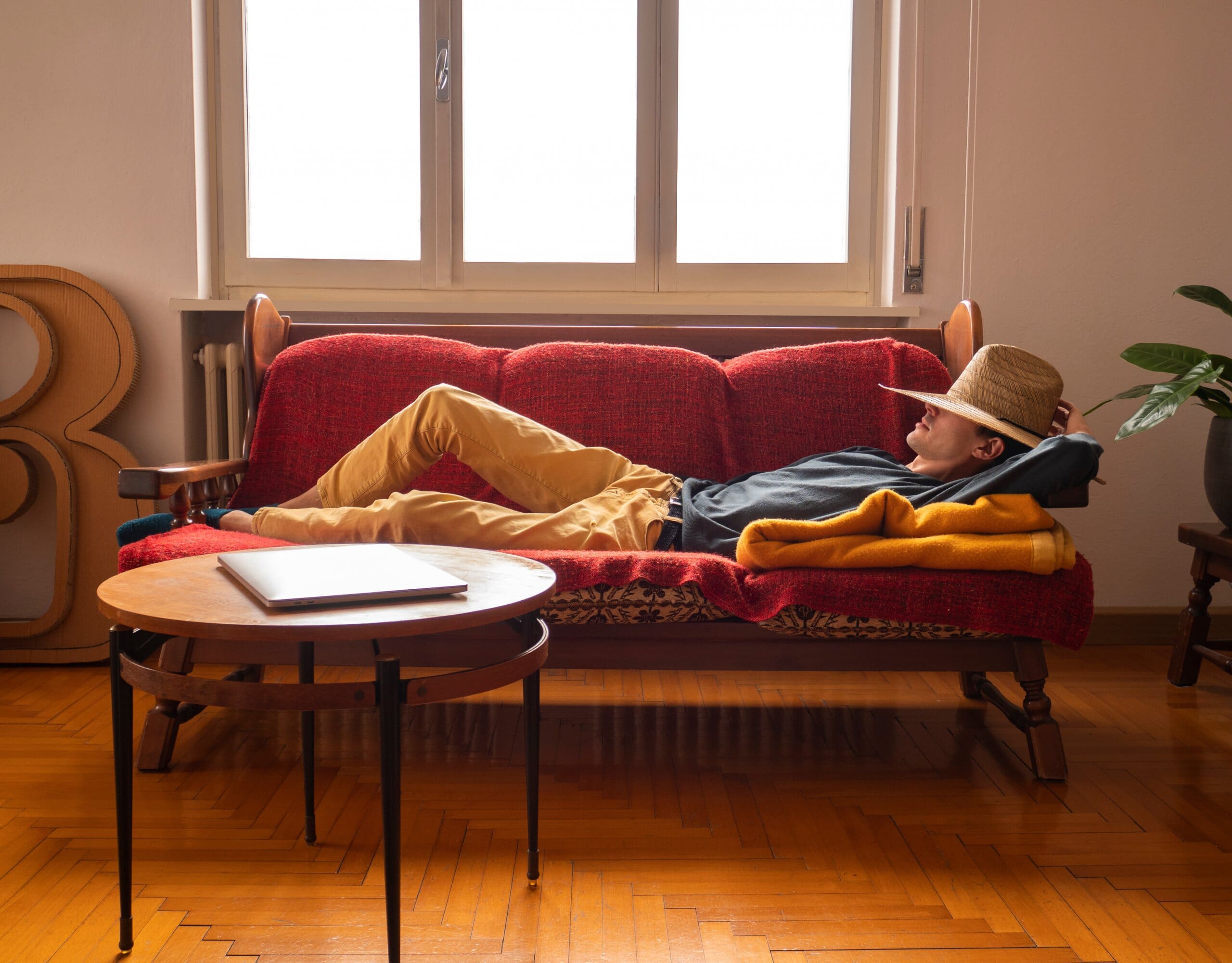 Man wearing hat over his face laying on a red sofa, having a nap.