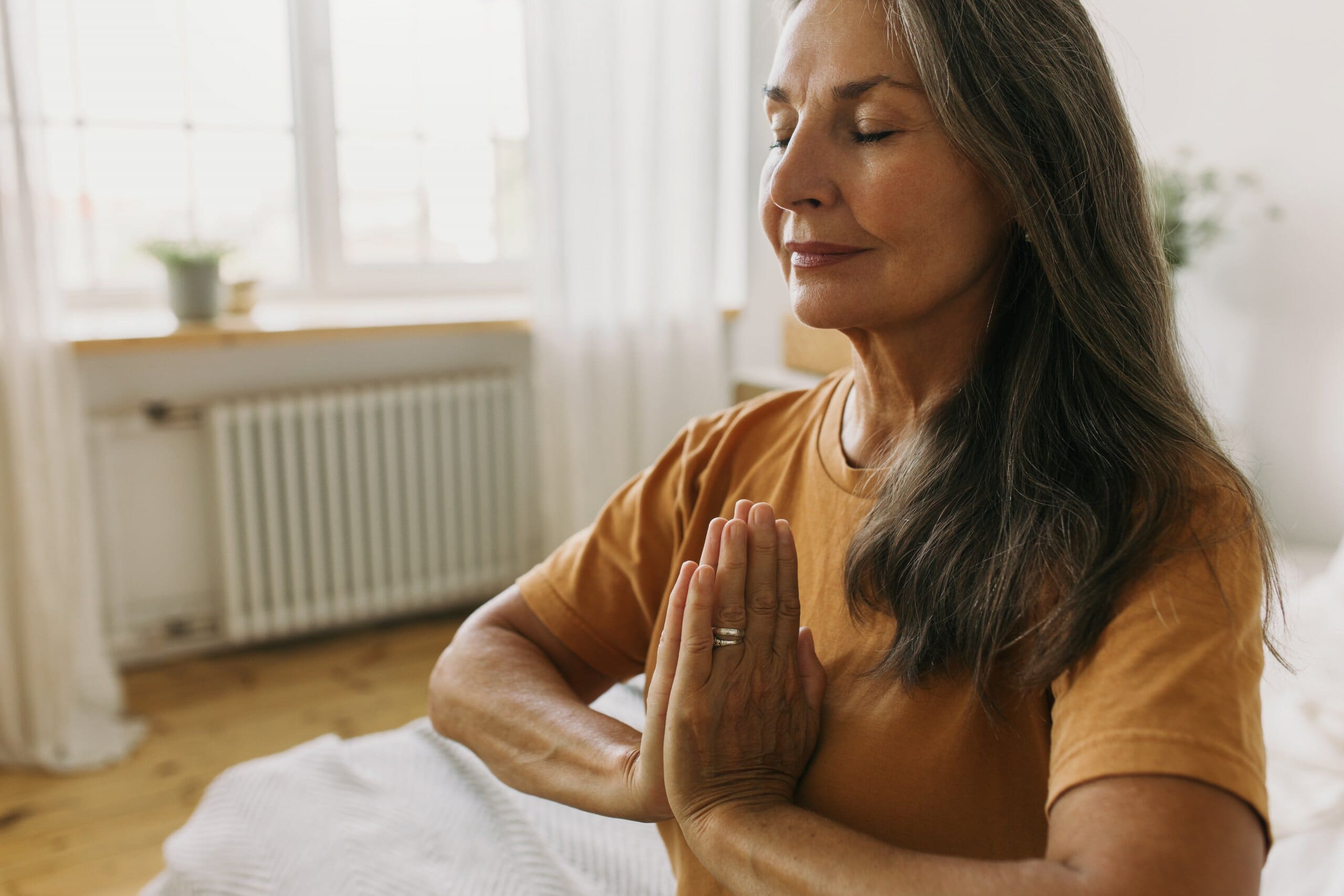 Middle aged lady sat at the end of a bed with her hands in a meditation pose.
