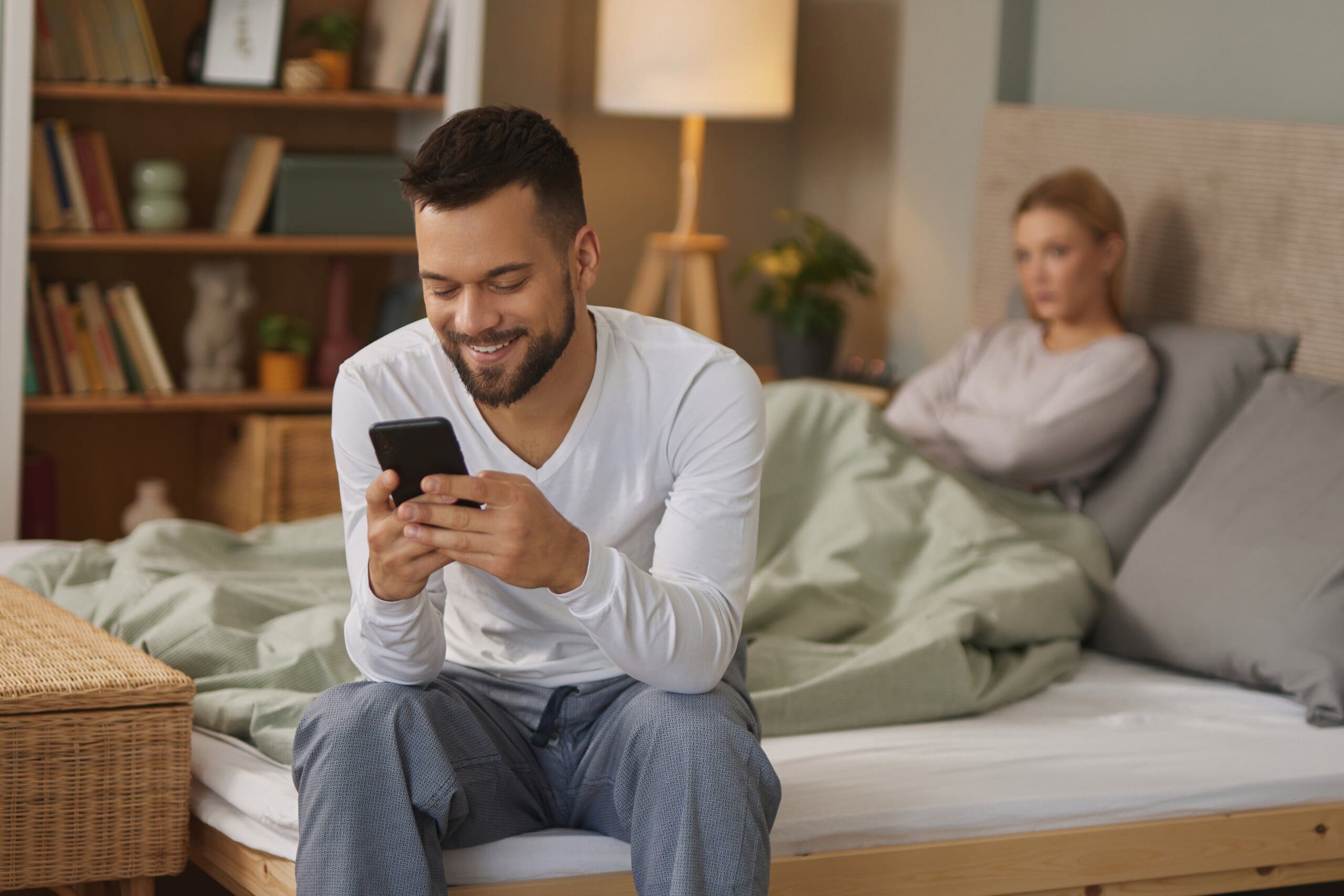 Man smiling at his phone sat on the end of the bed while woman looks upset laying in bed.