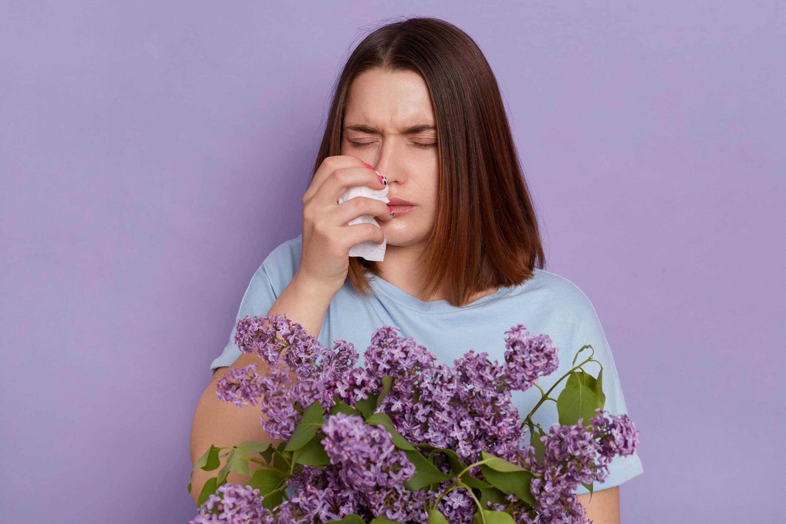 Woman suffering from Allergic Rhinitis sneezing as she holds a bunch of purple flowers.