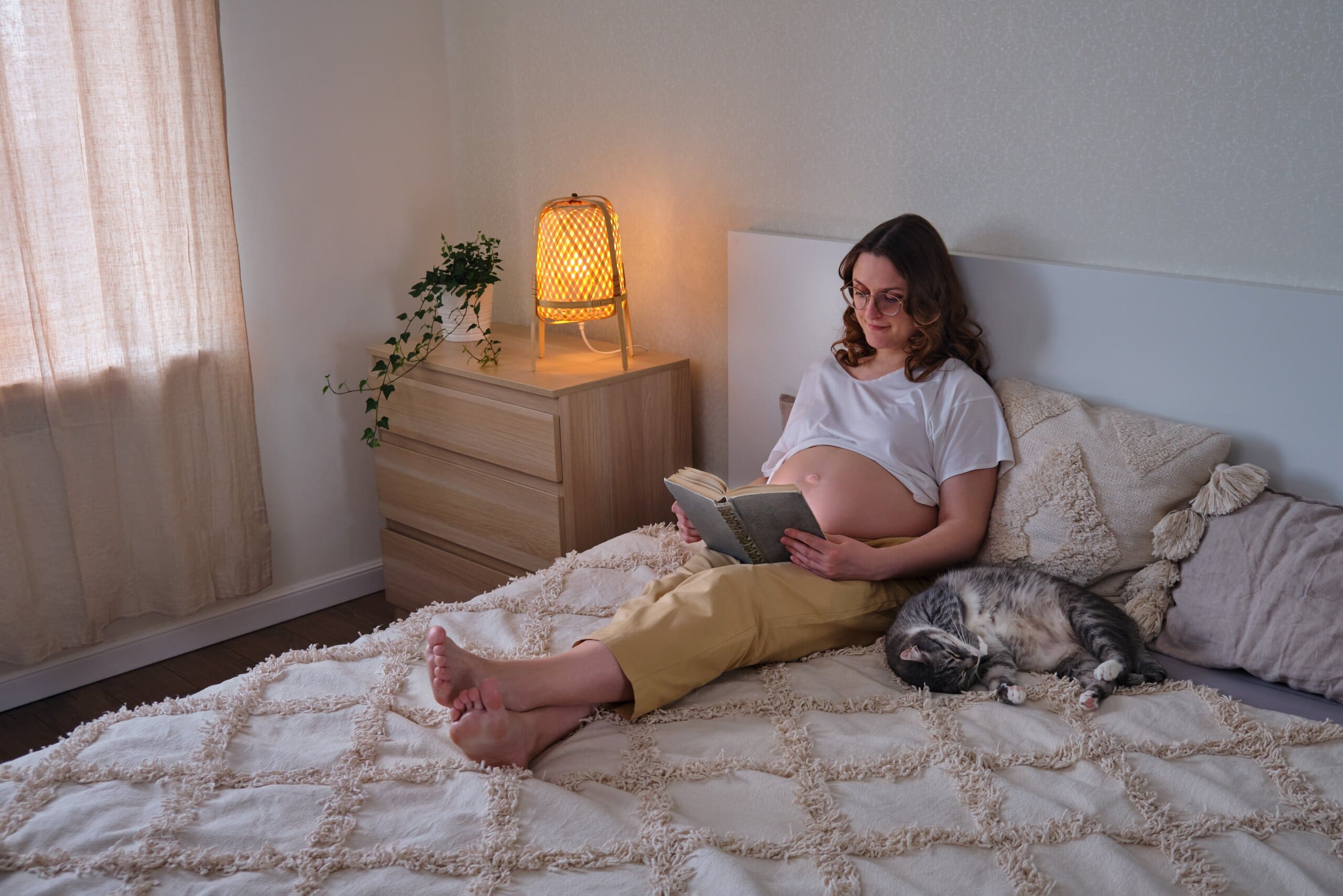 Pregnant lady reading a book in bed with a sleeping cat next to her.
