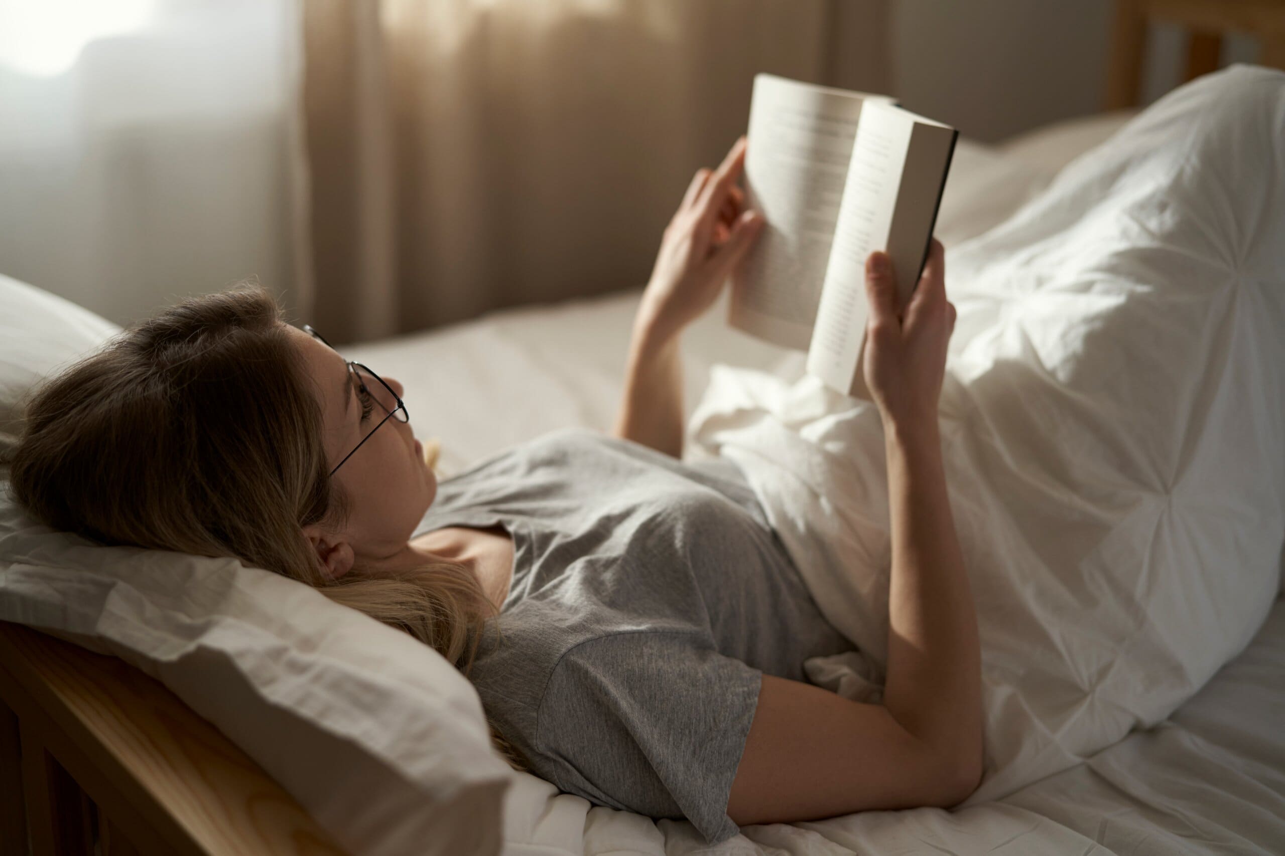 Woman with glasses laying in bed reading a book.