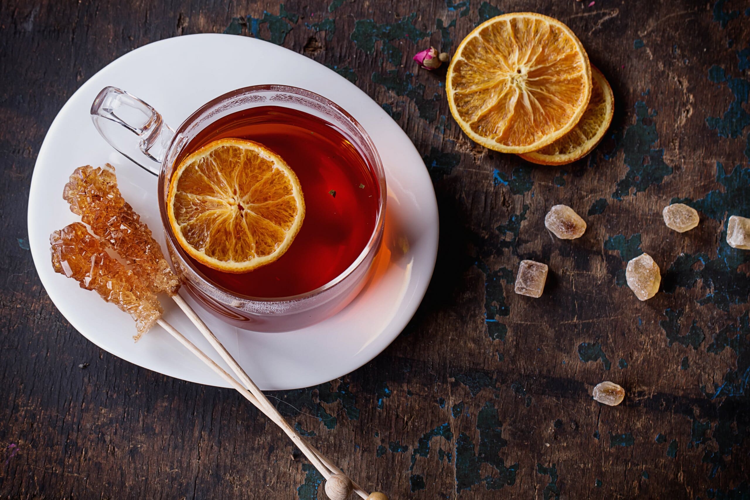 Mug of red bush tea surrounded by dried orange slices and candied sugar sticks on a saucer.