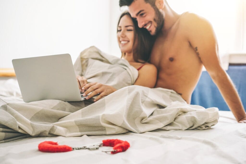 Couple in bed together on laptop, fluffy handcuffs laying next to them, shopping for the best mattresses for sex.