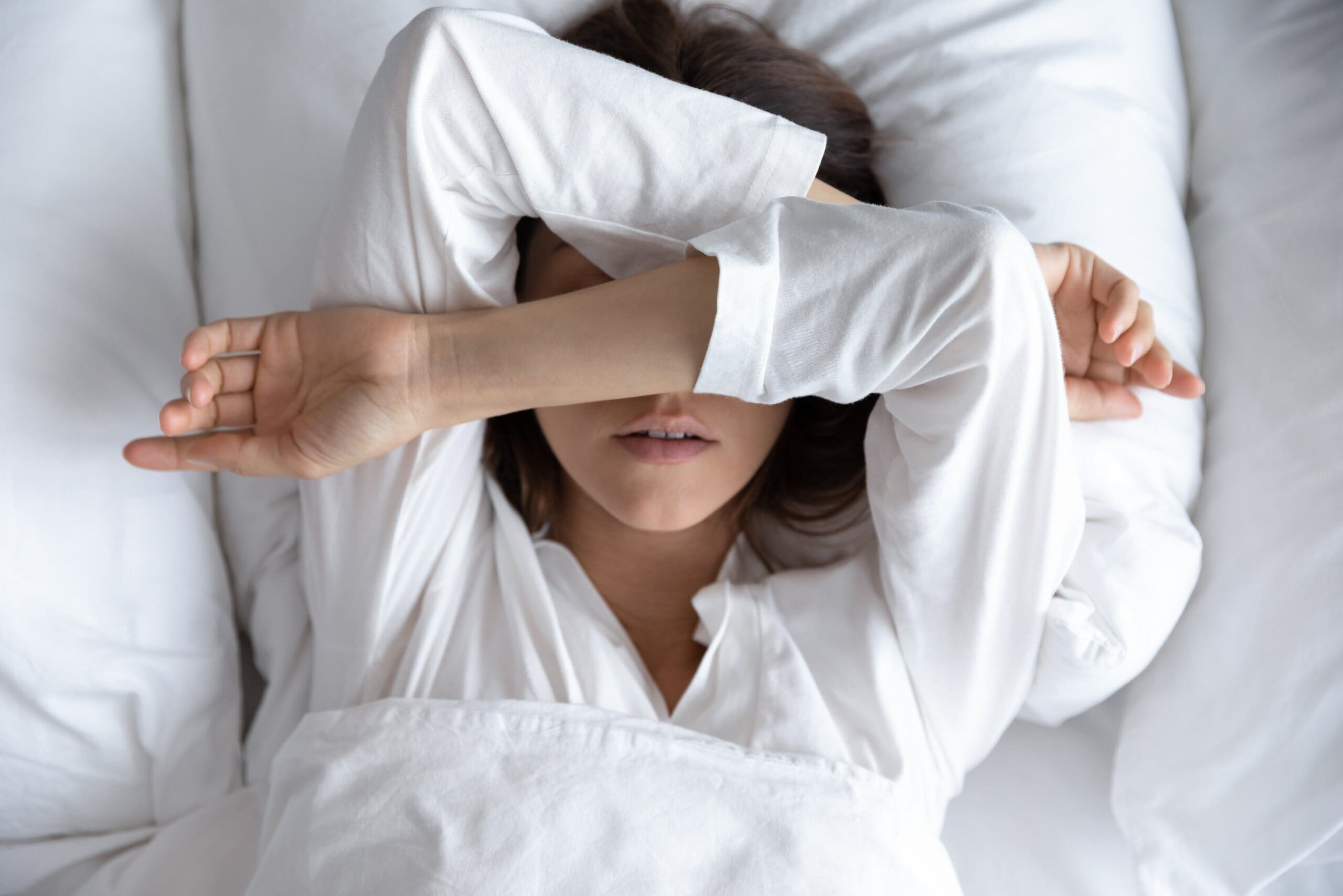 Head shot above top view exhausted young woman lying in bed under duvet, covering eyes with hands, suffering from sleep anxiety and insomnia.