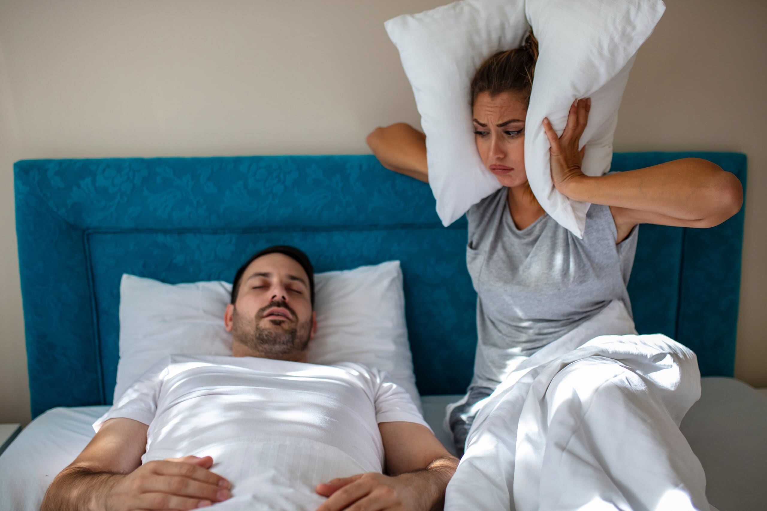 Man snoring or sleep talking, waking his partner who is holding a pillow to her ears sat up in bed next to him.