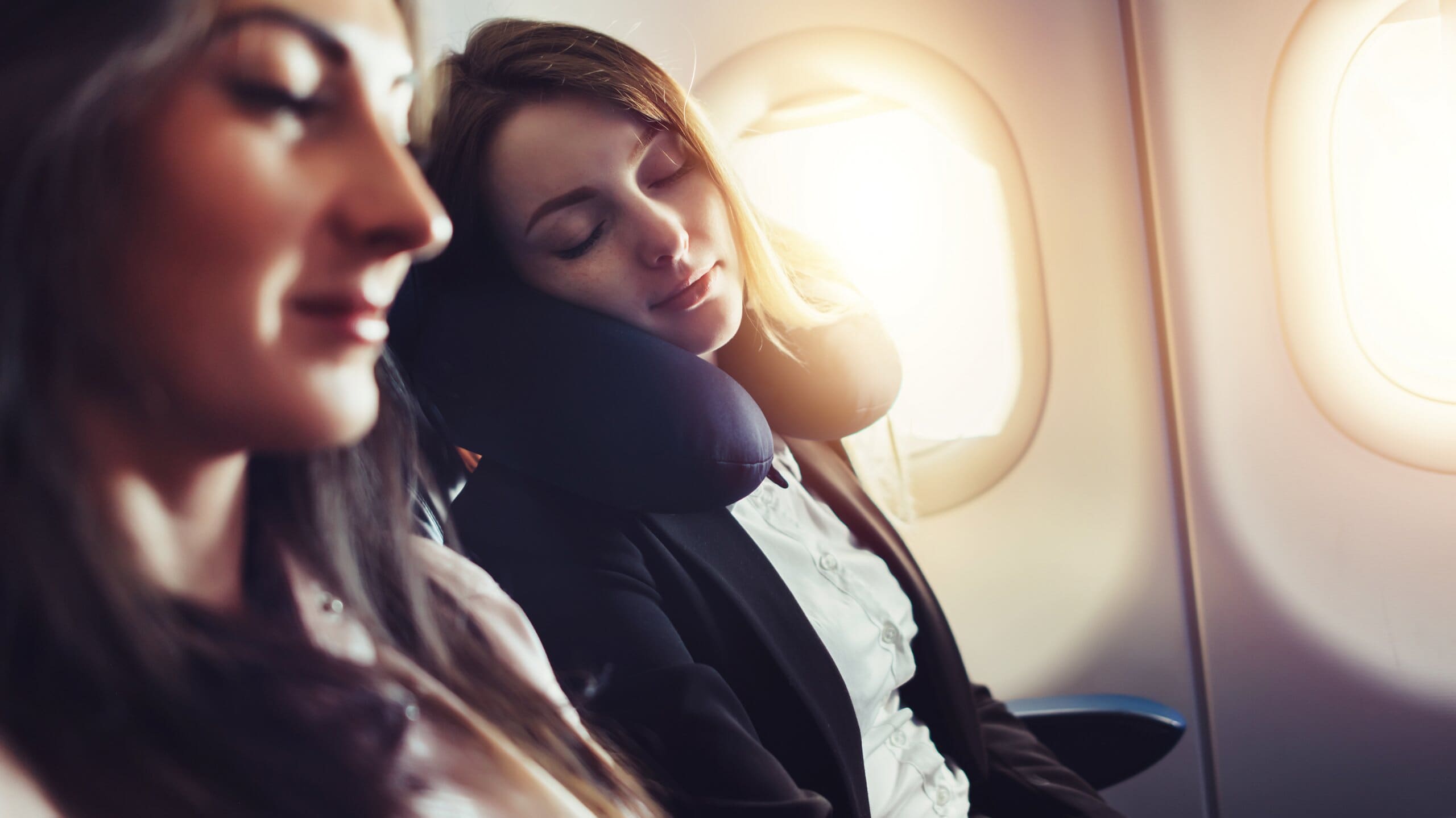 Two women on a plane, one sleeping with a memory foam travel pillow around her neck.