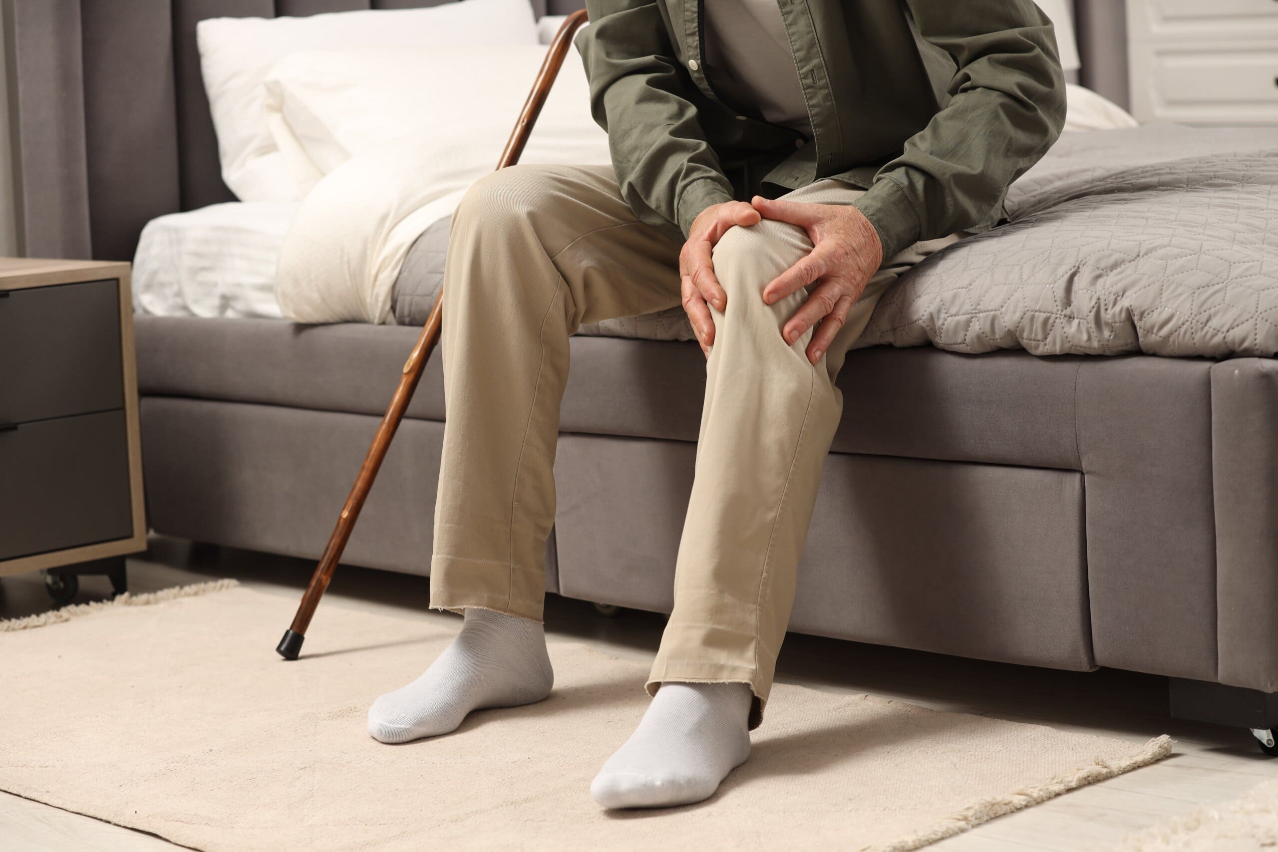 Cropped image of an elderly man holding his knee sitting on the edge of his bed.