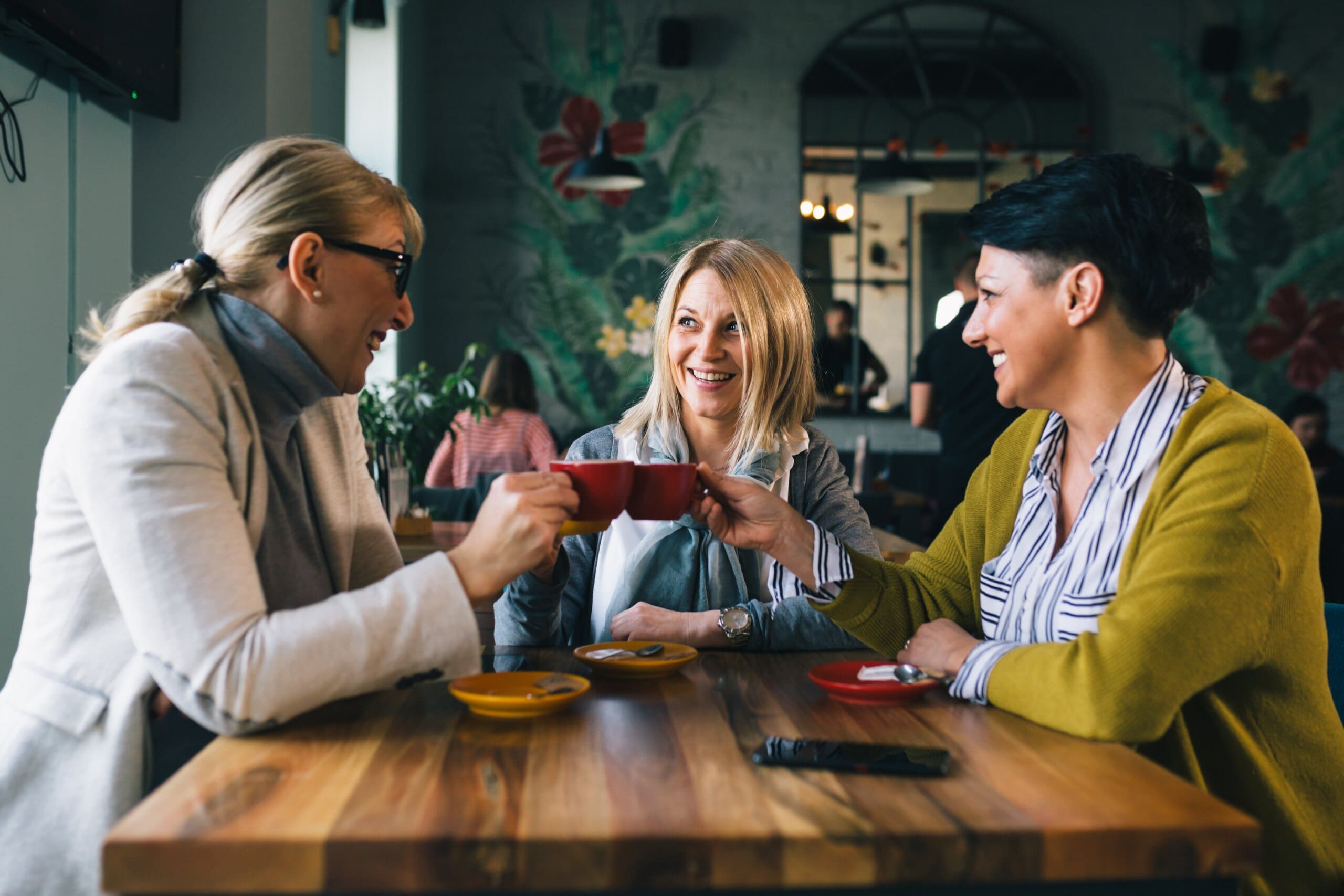 Three middle aged women laughing chatting drinking coffee in a cafe.