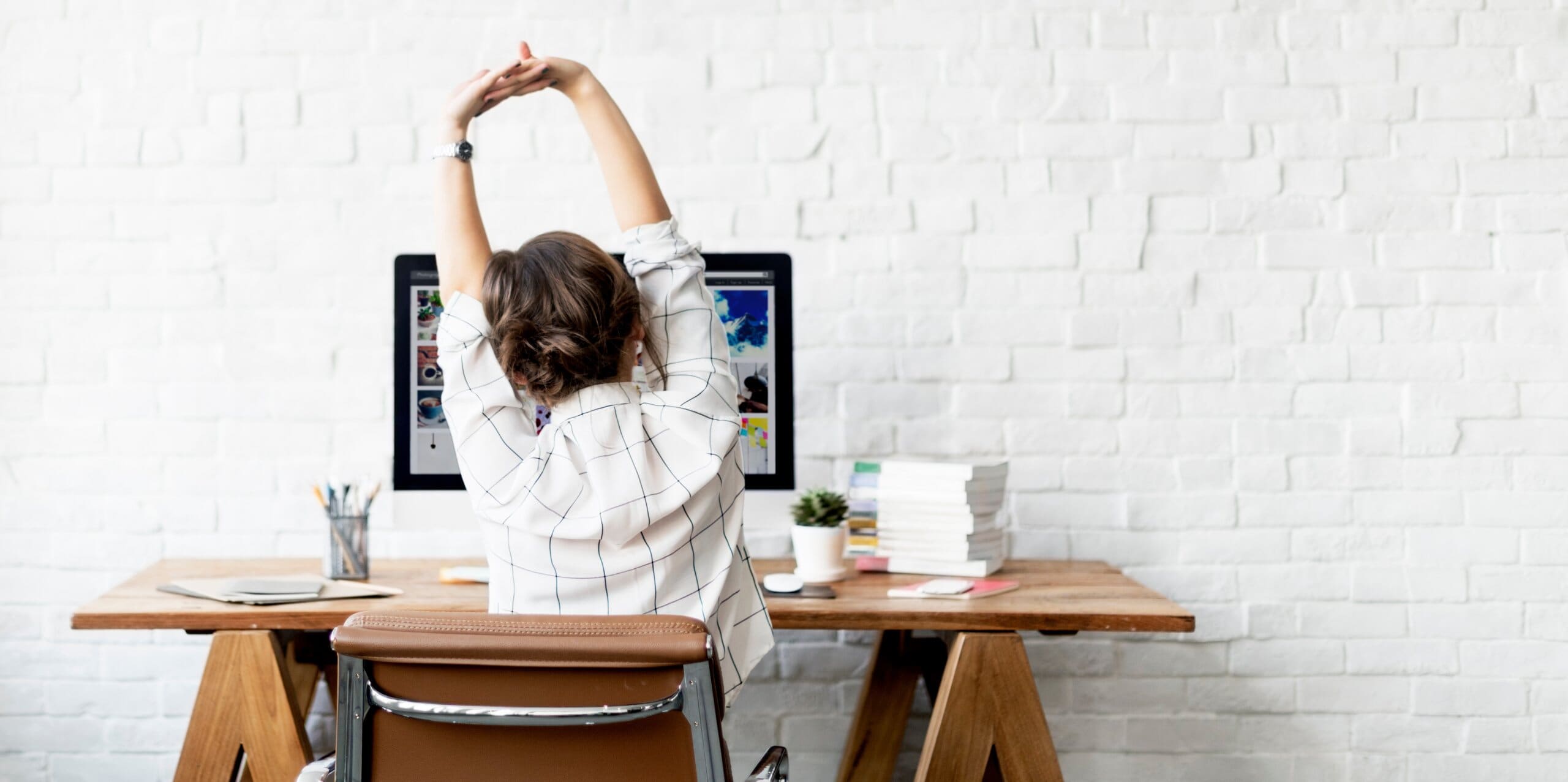 Shot of a woman from behind stretching her arms above her head at her work desk.