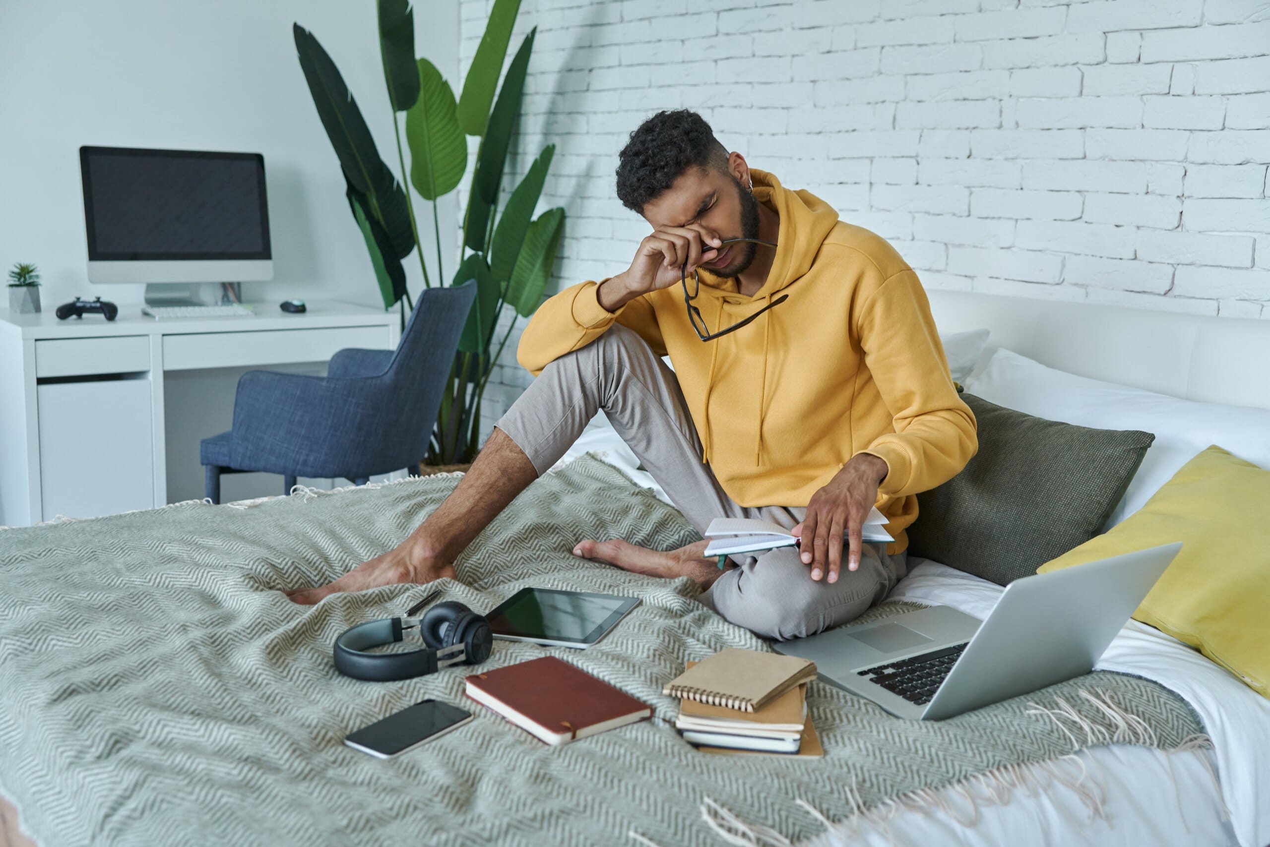 Male student sitting on bed looking stressed, surrounded by laptop and notebooks.