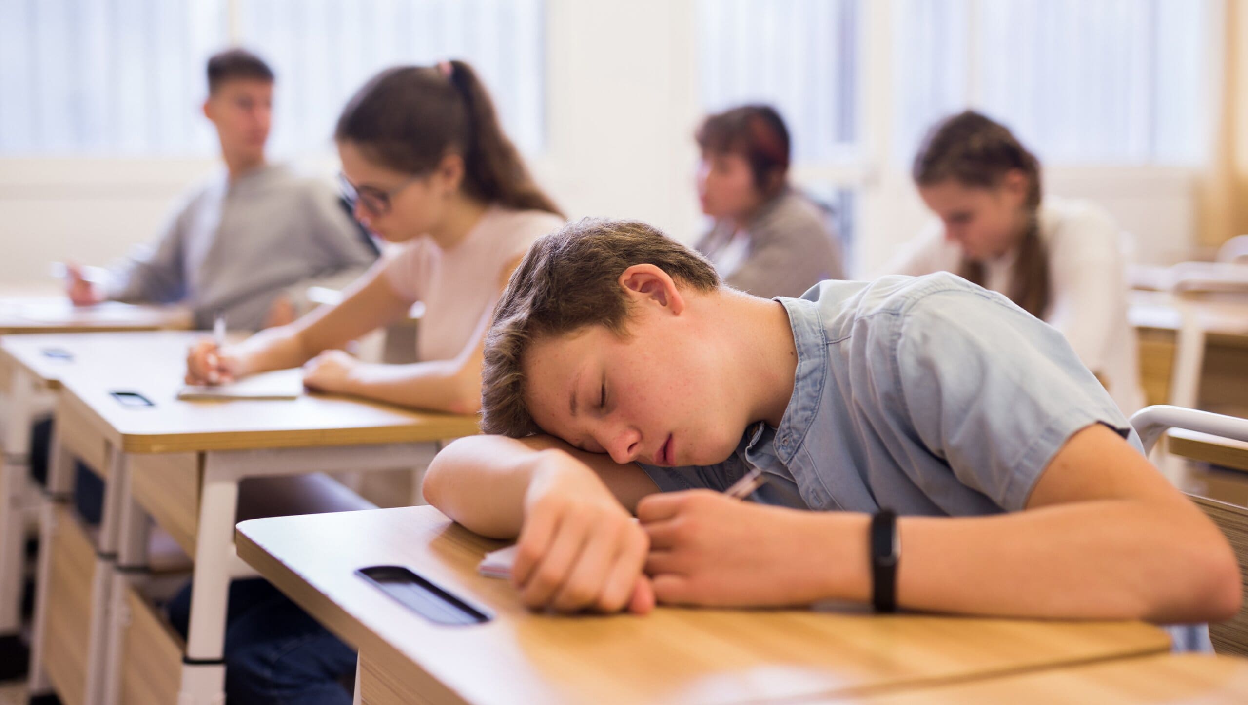 Tired teenage boy who needs more sleep, asleep at his school desk, with blurred classmates in the background.